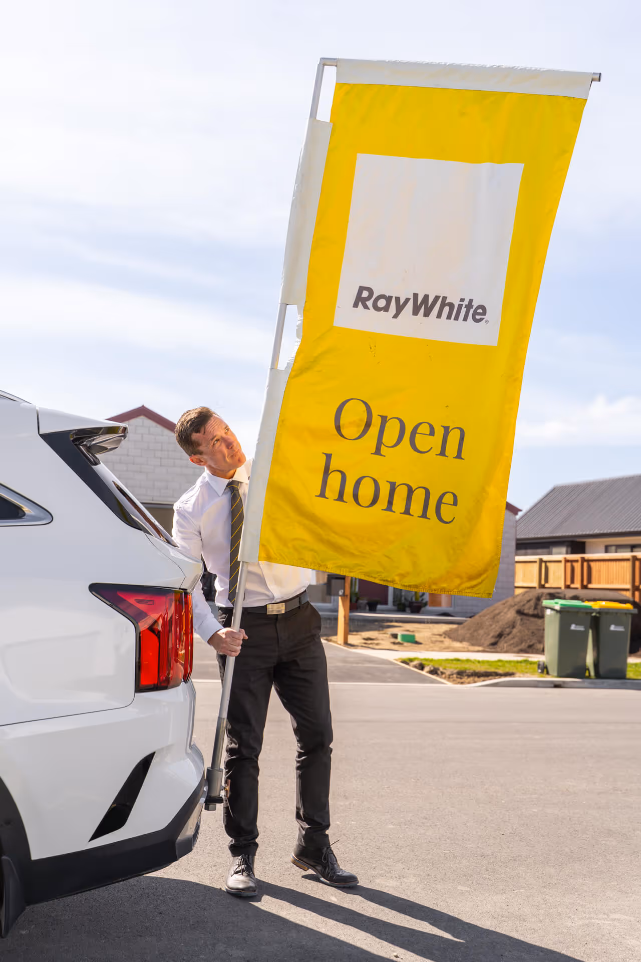Man in business attire holding a yellow RayWhite Open home flag next to a white car in a suburban neighborhood.