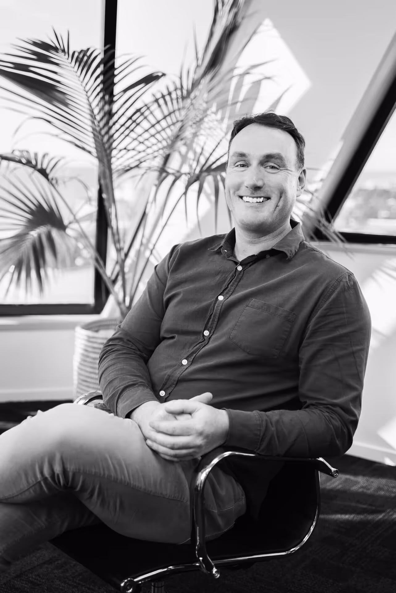 Smiling man sitting cross-legged in an office chair with plants and large windows in the background.