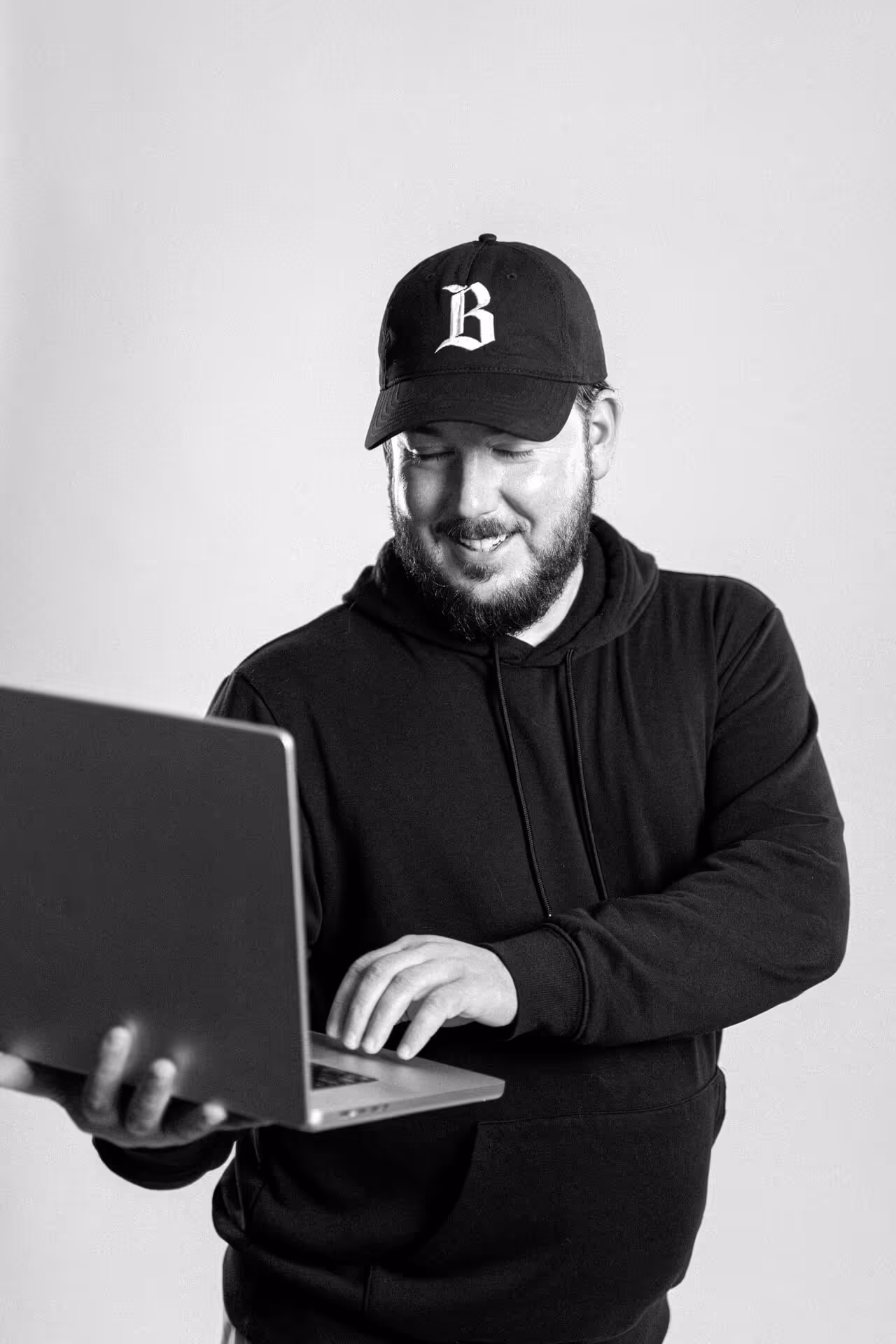Bearded man wearing a black cap and hoodie smiling while holding and using a laptop.