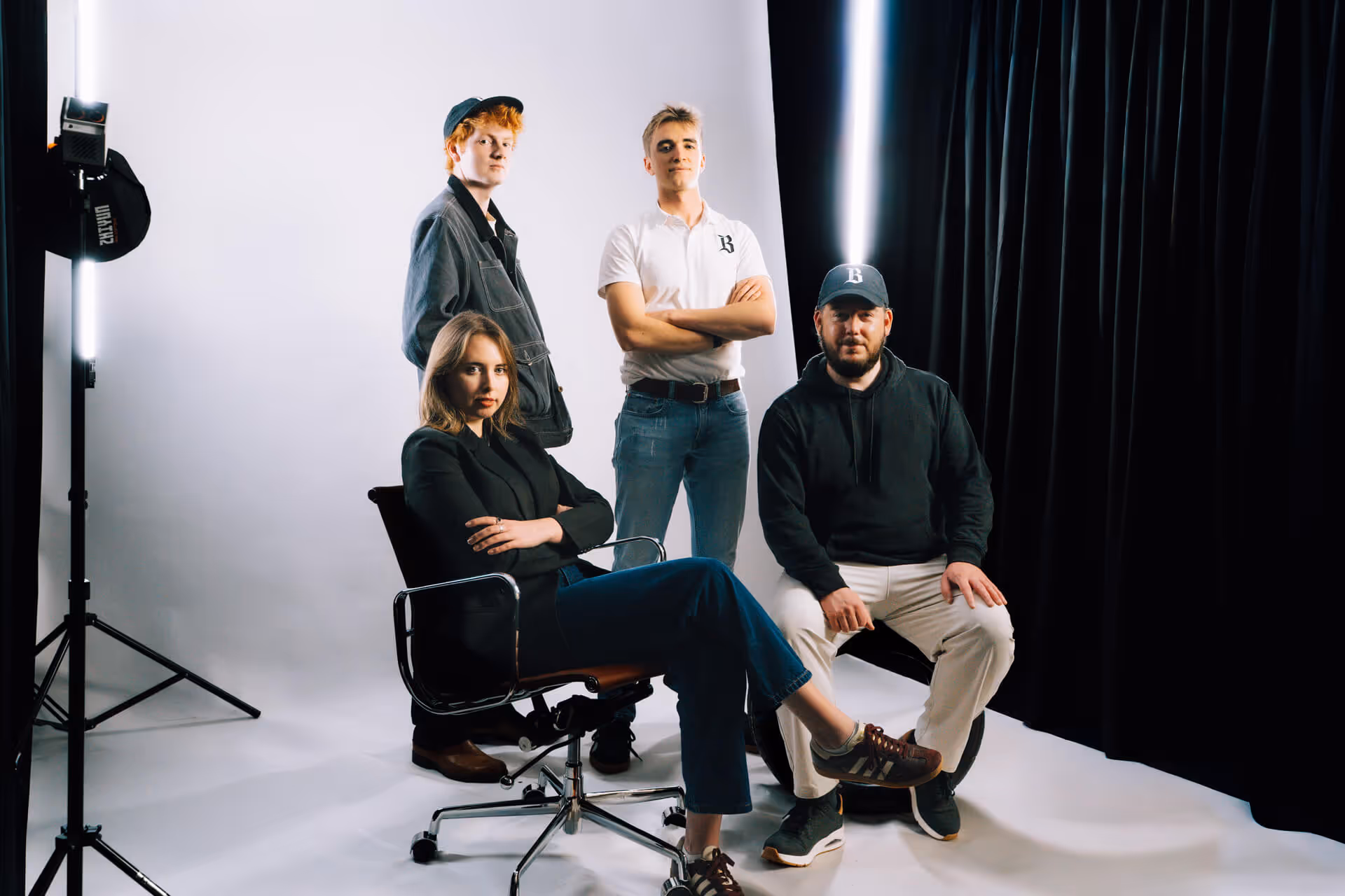 Four young adults in a studio: two sitting on chairs and two standing, with white and black backgrounds and studio lighting.