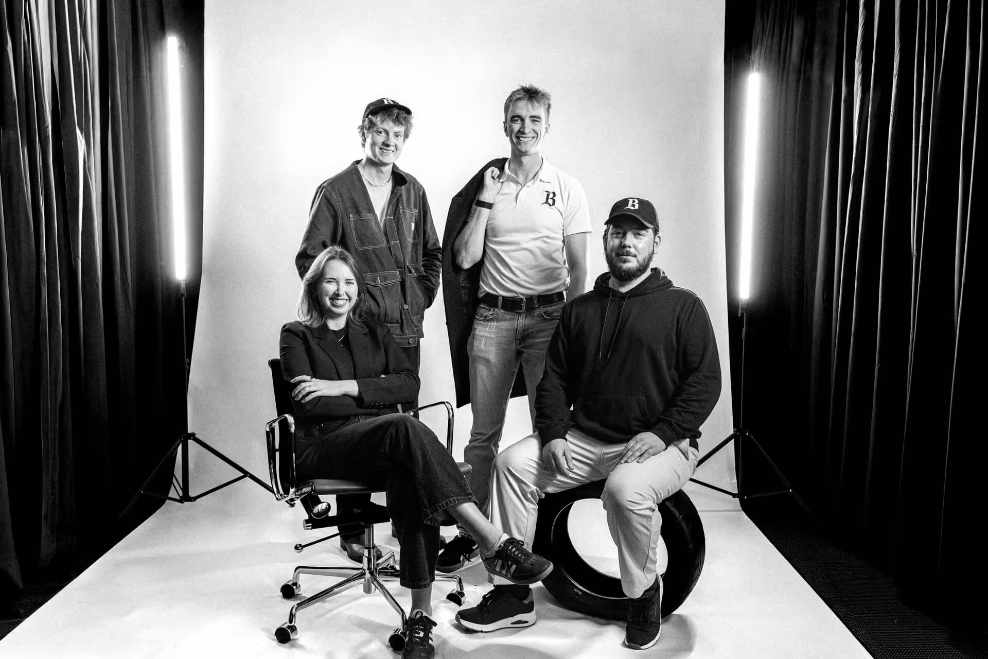 Black and white photo of four smiling people in a studio setting with two vertical lights and dark curtains; two are seated and two are standing.