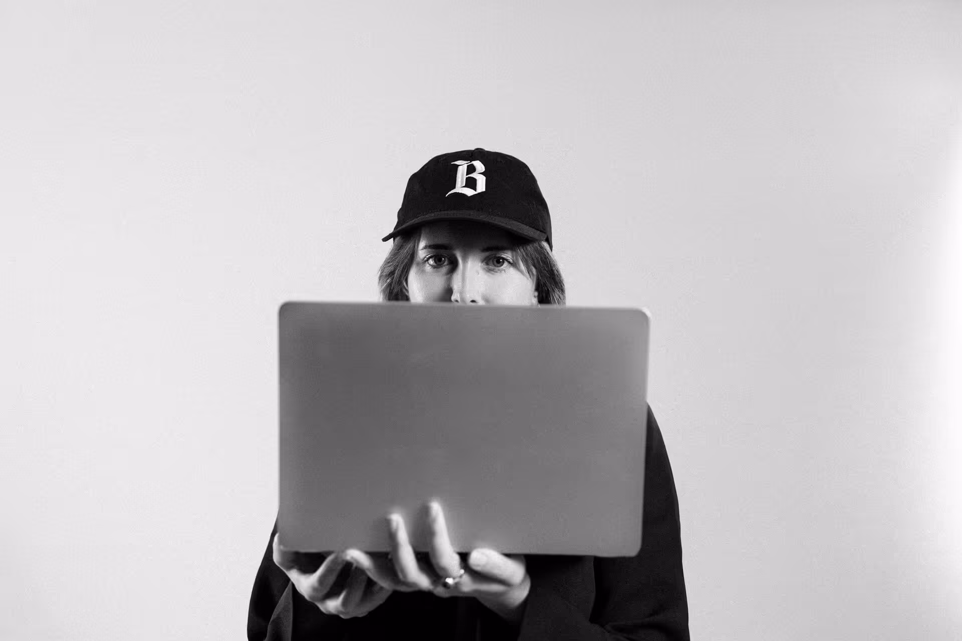 Person wearing a black cap holding a laptop in front of their face, looking directly ahead.