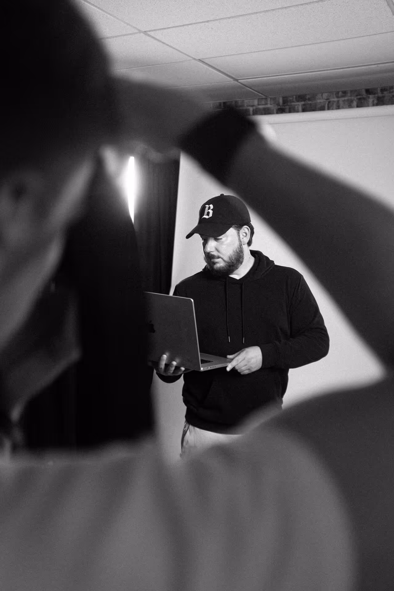 Man wearing a cap and hoodie looking at a laptop, with a blurred person holding a camera in the foreground.