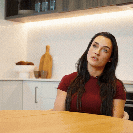 Woman with long dark hair wearing a red shirt sitting at a wooden table in a modern kitchen.