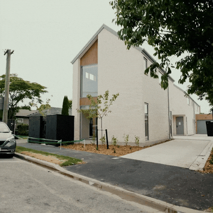 Modern two-story house with beige brick exterior, large front window, driveway, and small trees in front yard.