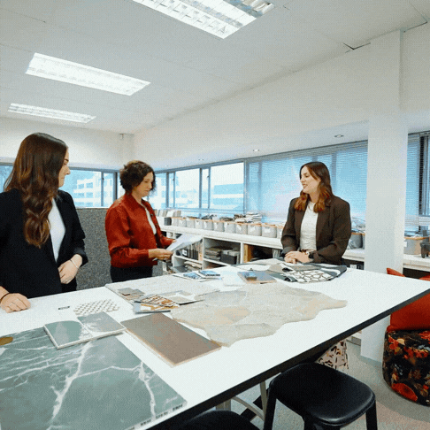 Three women in a modern office discussing samples and materials spread on a white table near large windows.
