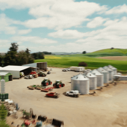 Aerial view of a rural farm with silos, tractors, farming equipment, and green fields under a partly cloudy sky.
