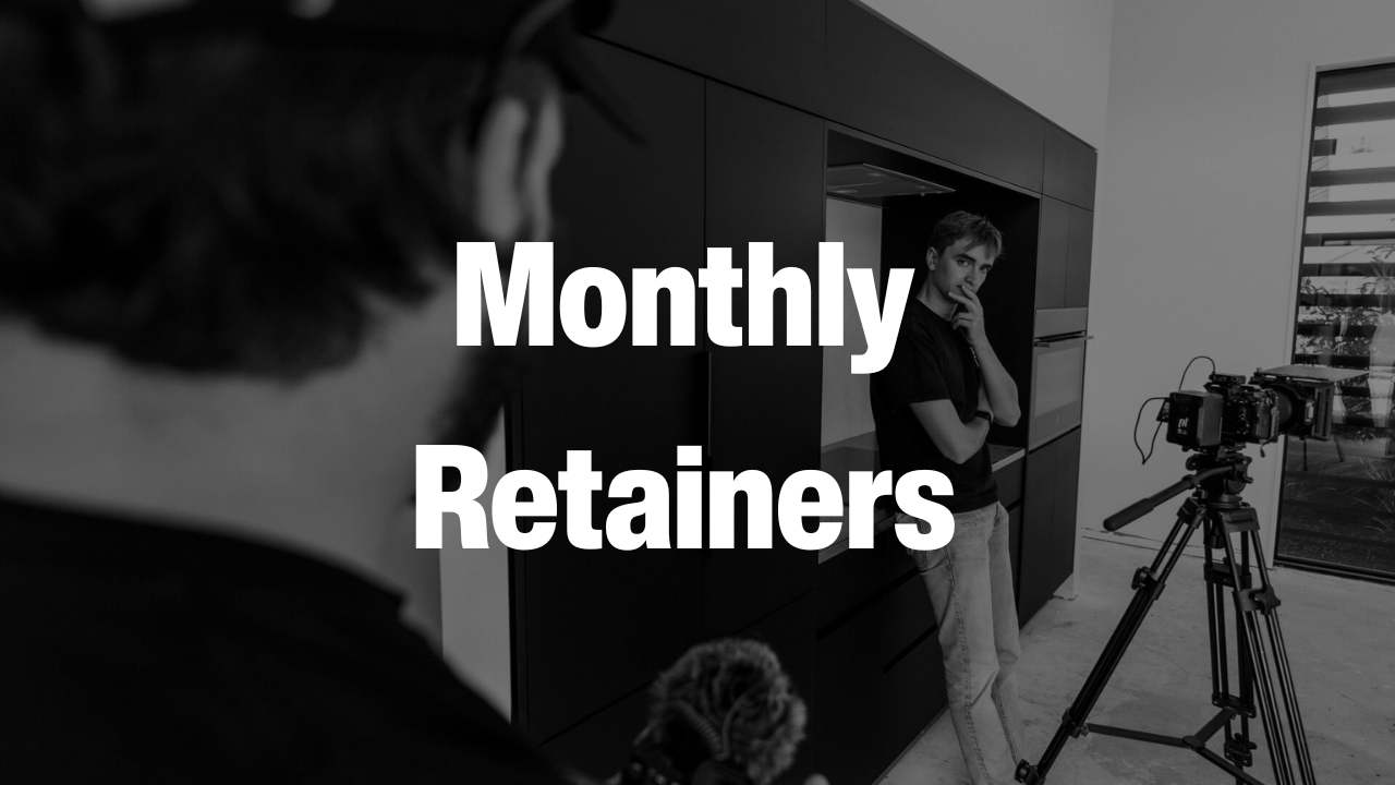Black and white photo of a man standing thoughtfully near a camera on a tripod inside a modern kitchen with another person partially visible in the foreground.
