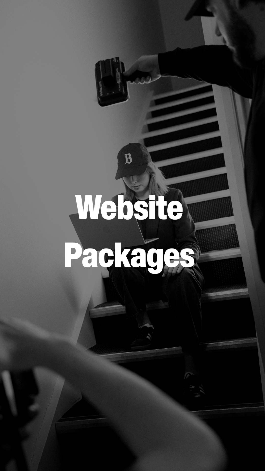 A person wearing a cap sits on carpeted stairs using a laptop while another person points a handheld camera above them.