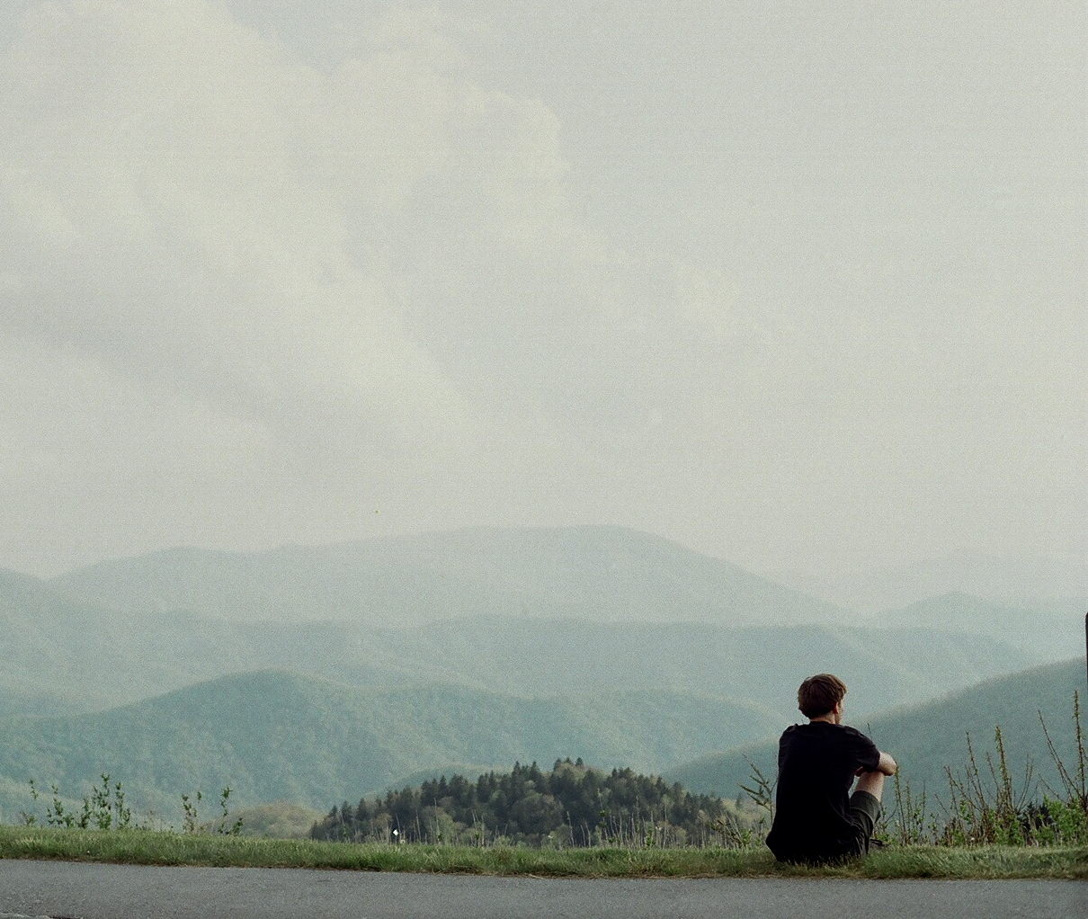 Man sits meditatively at a mountain overlook gazing out towards the horizon. 