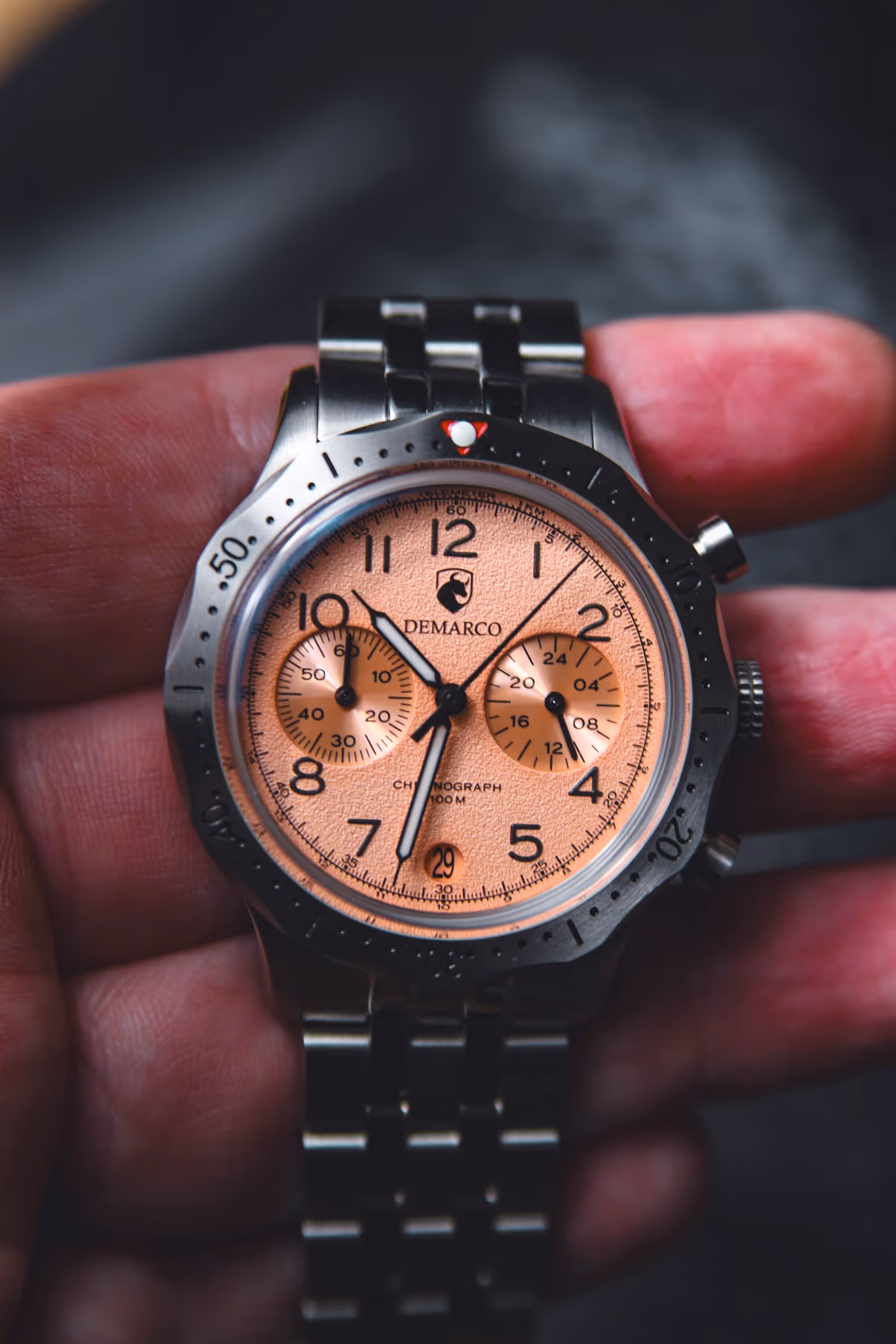 A DeMarco chronograph with a salmon dial held in the hand on its black steel bracelet