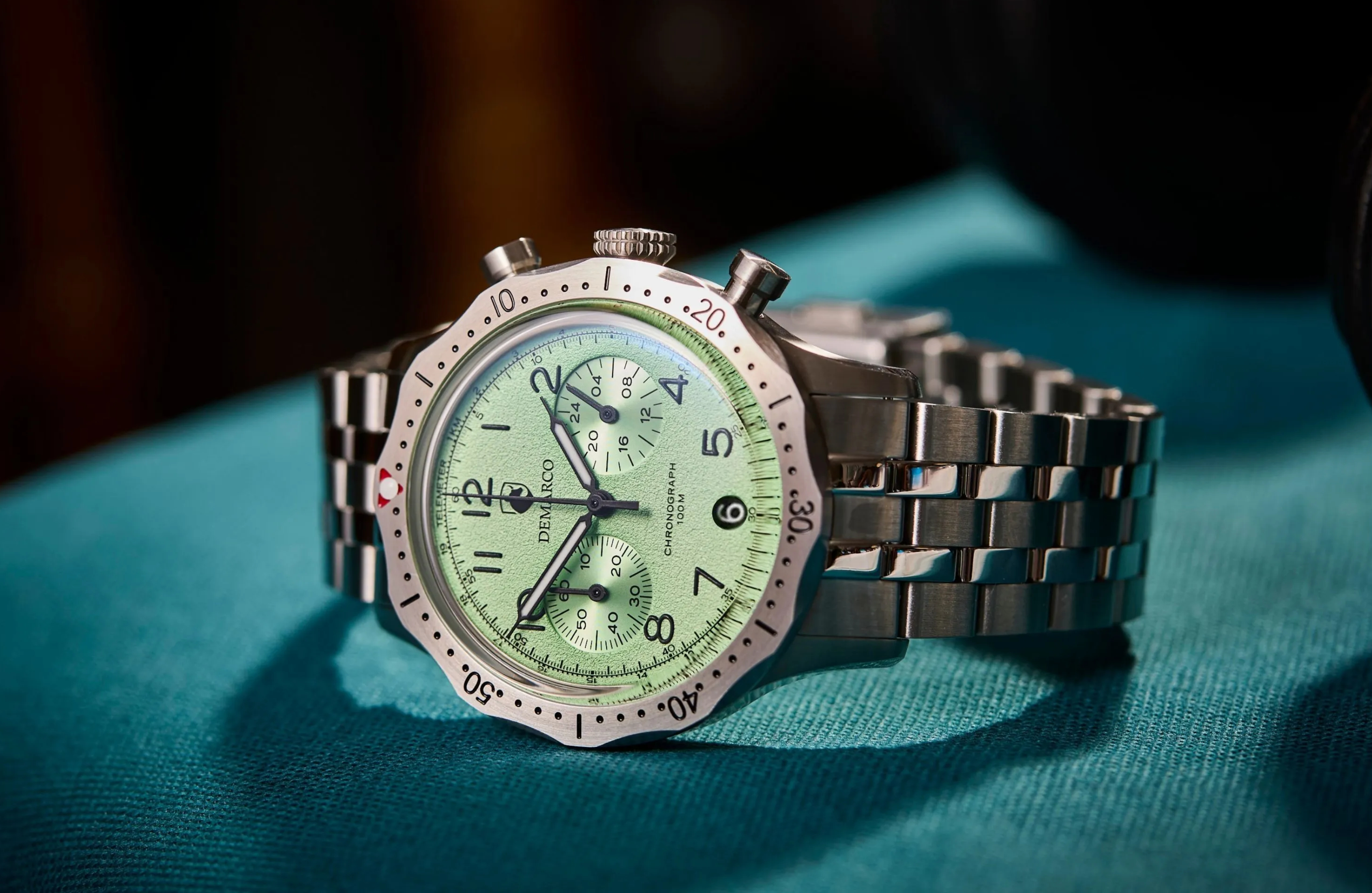 A DeMarco chronograph with a pale green dial on a steel bracelet, lit against a teal background