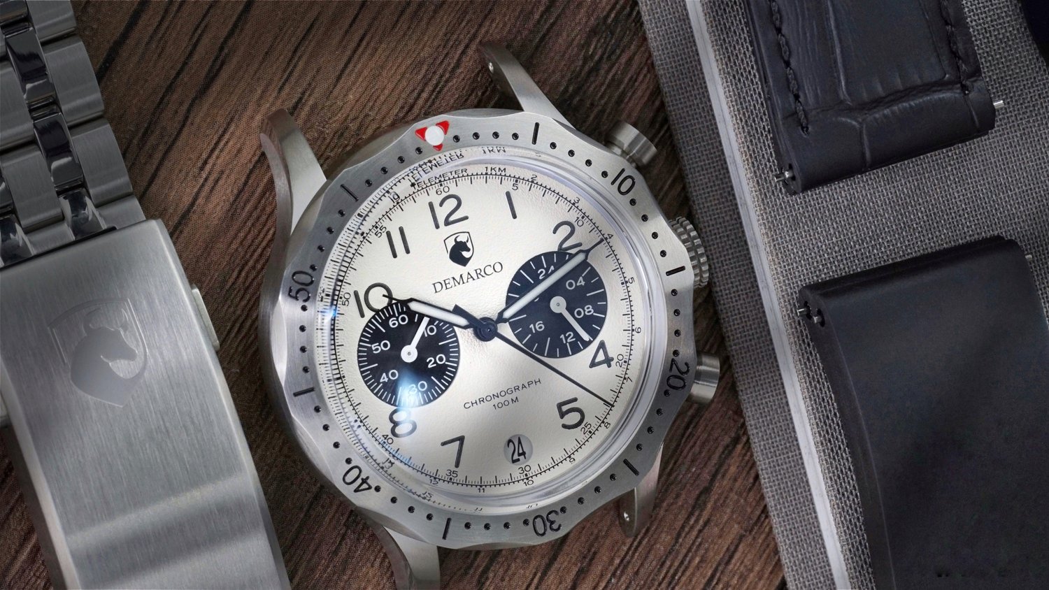 A DeMarco chronograph with a white dial on a wooden surface, shown with its steel bracelet and a folded leather strap