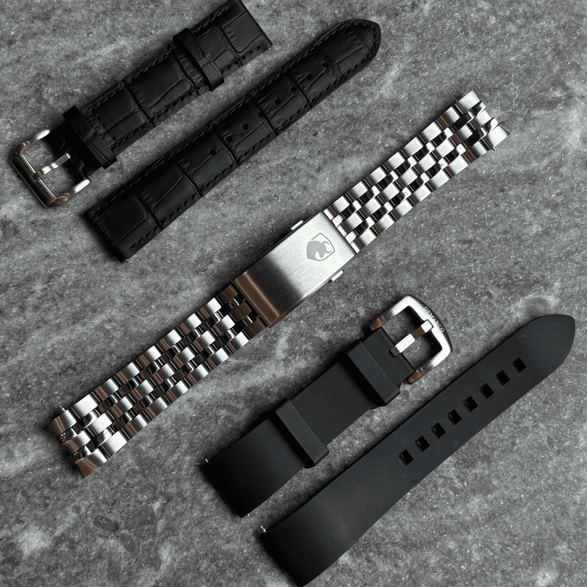Four DeMarco straps laid out on stone: black leather, a steel bracelet with signed clasp, brown leather and black rubber