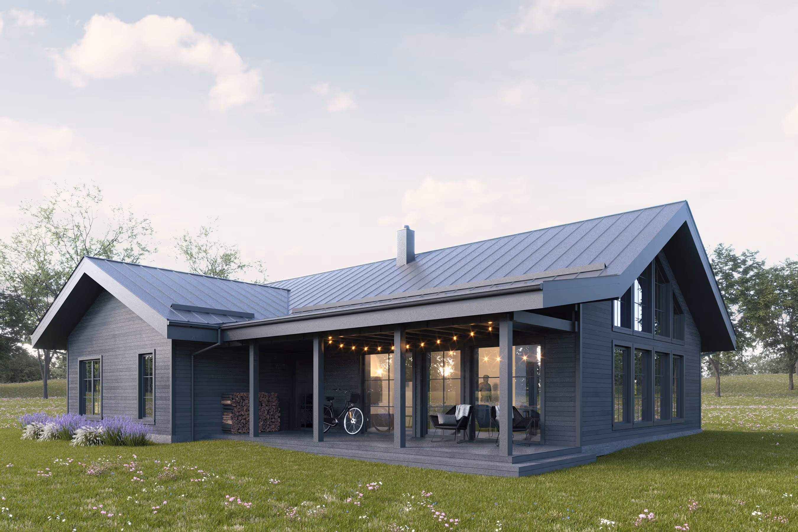 Modern dark gray wooden house with large windows, covered porch with seating and stacked firewood, set in a grassy field with trees.