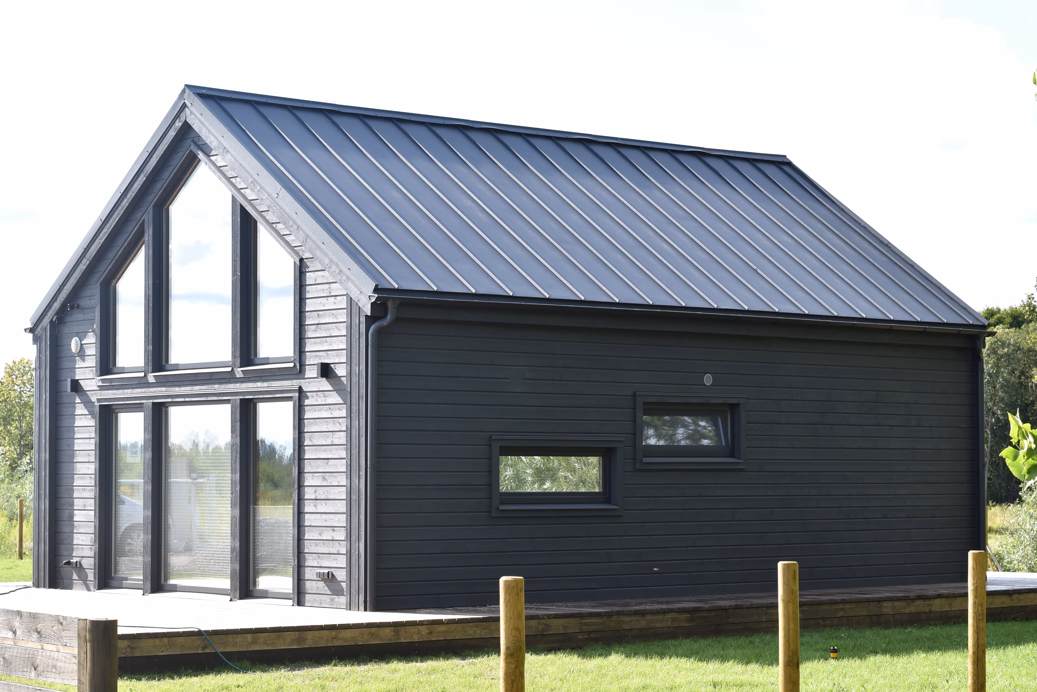 Modern black wooden house with large triangular windows and a metal roof, surrounded by grass and wooden posts.