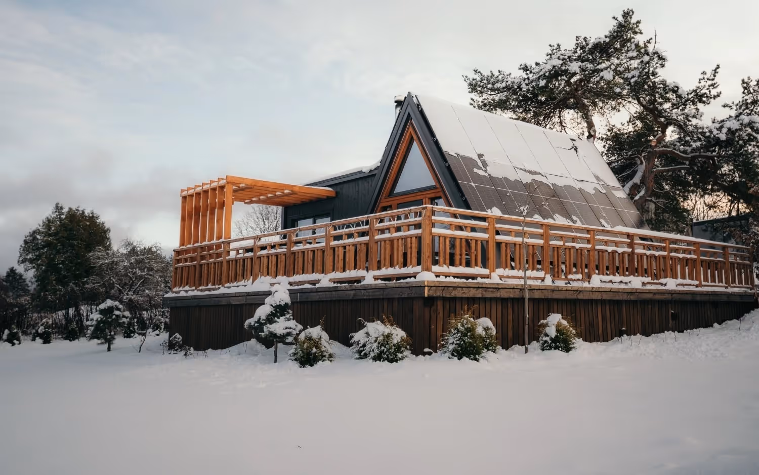 A-frame cabin with solar panels on roof and wooden deck covered in snow in a snowy landscape.