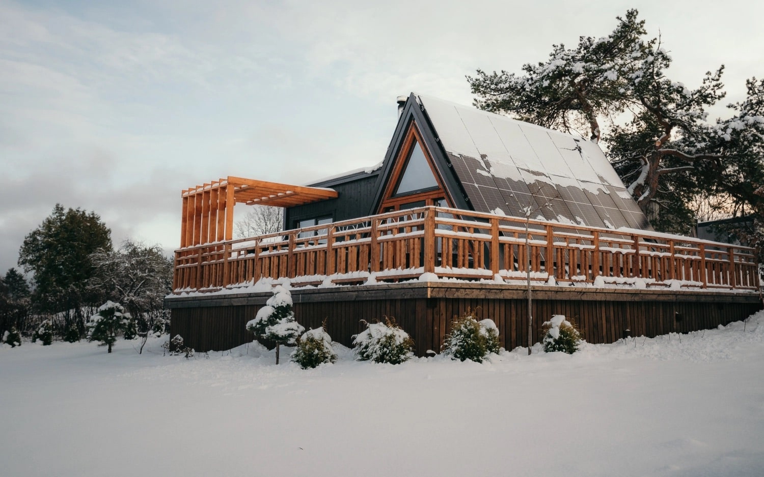 A-frame cabin with solar panels on roof and wooden deck covered in snow in a snowy landscape.