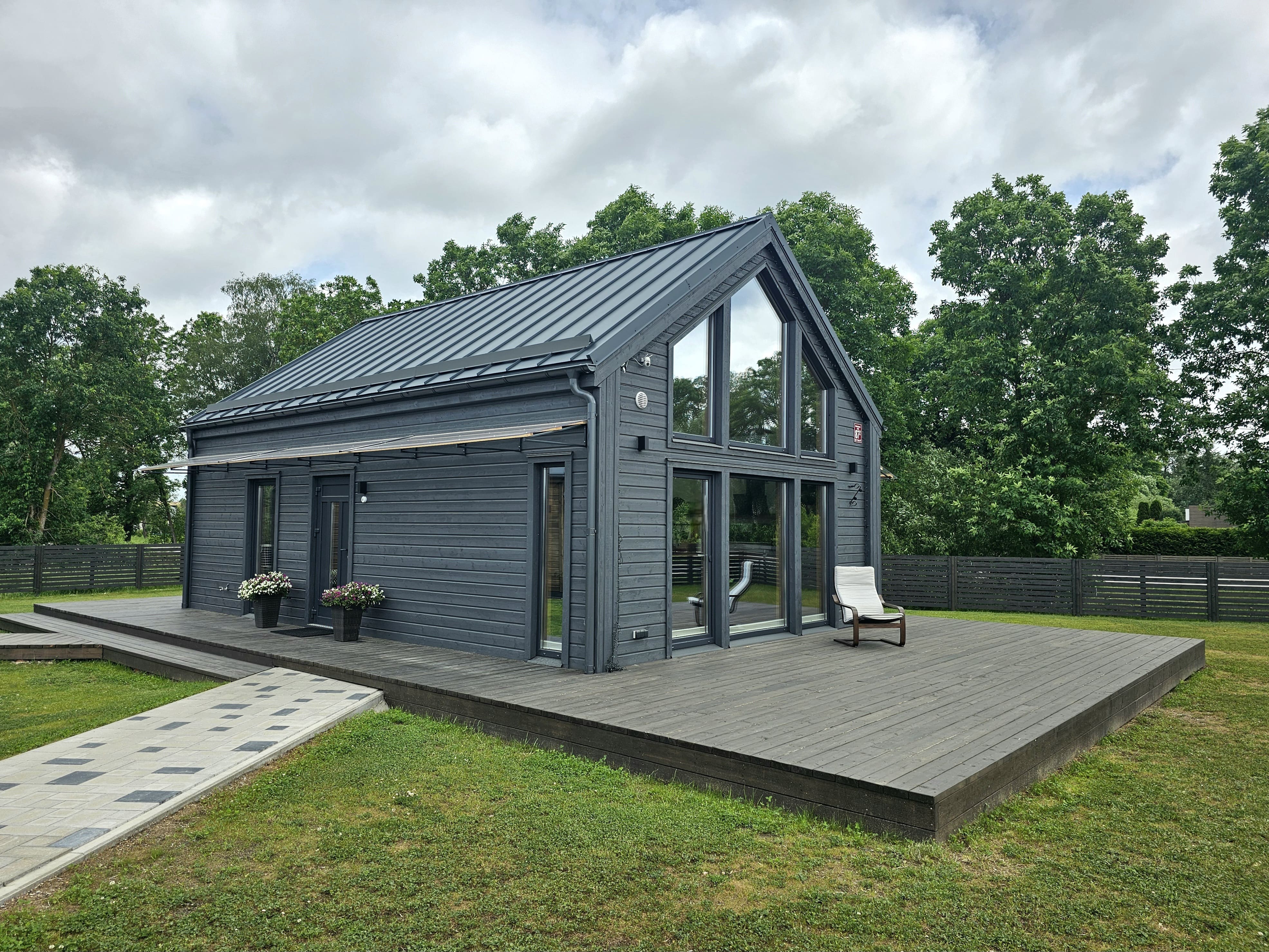 Modern small dark gray wooden house with large windows and a wooden deck with a single chair, surrounded by green grass and trees under a cloudy sky.