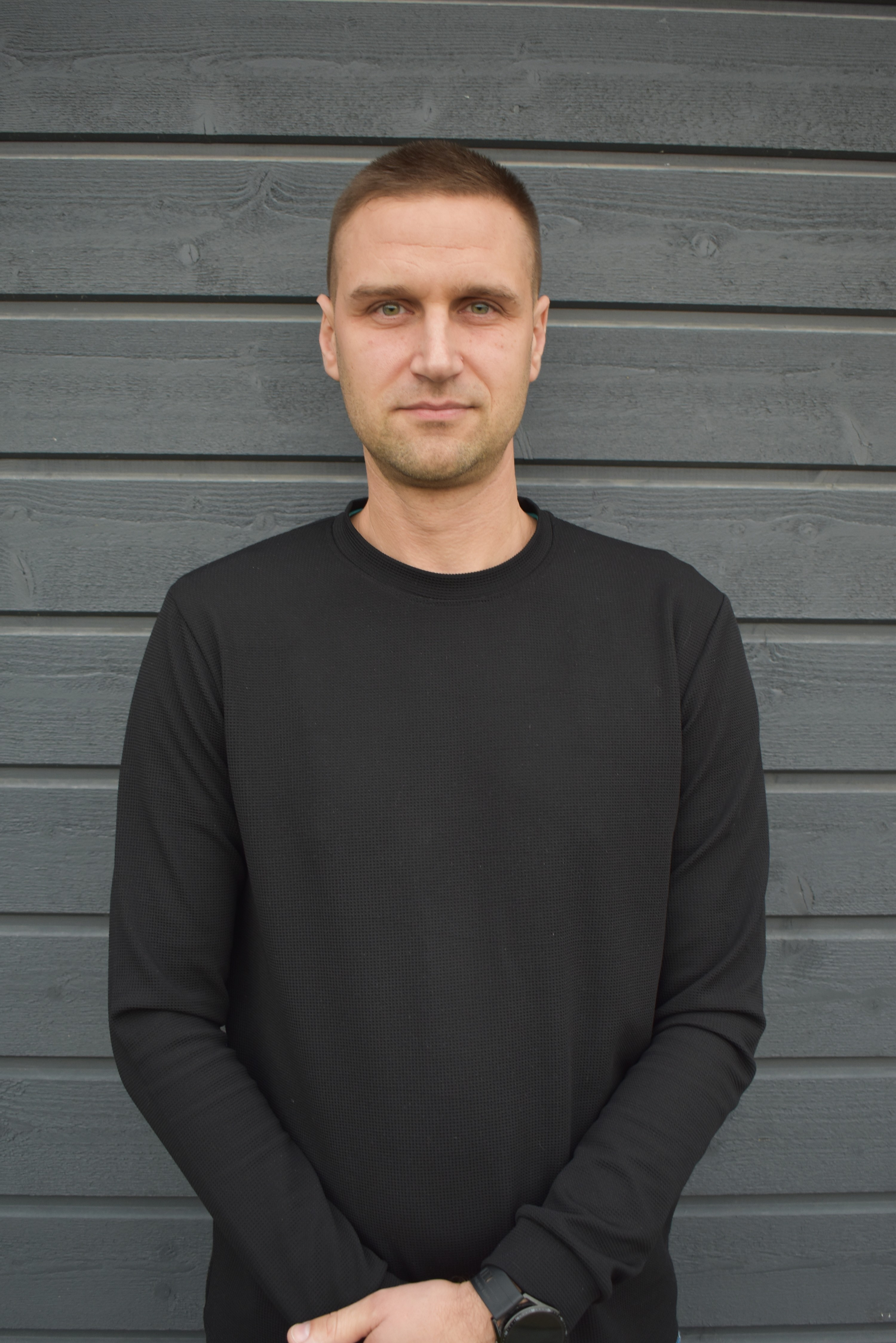 Portrait of a man with short brown hair wearing a black long-sleeve shirt standing against a gray wooden wall.