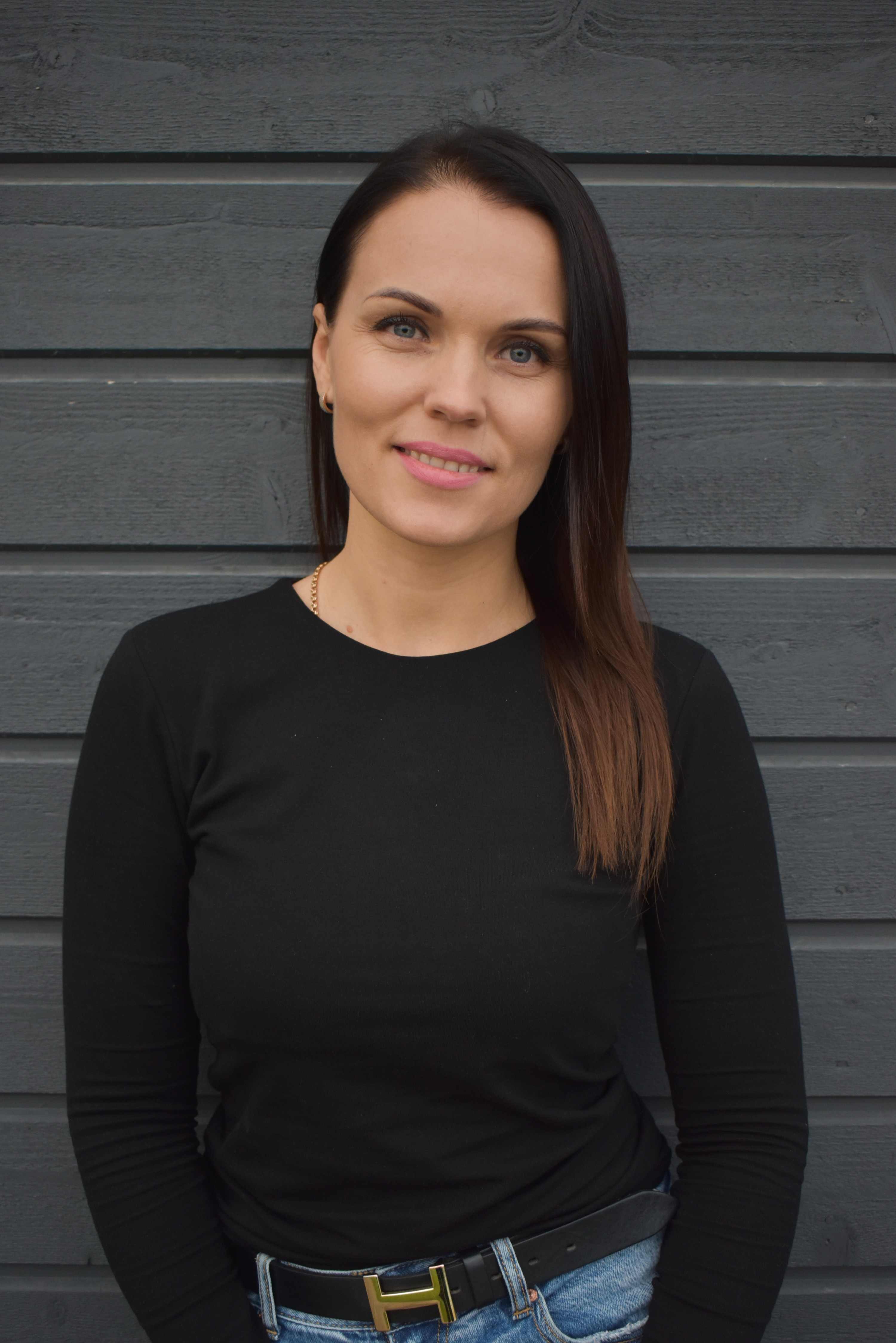 Woman with long dark hair wearing a black long-sleeve shirt and blue jeans, standing in front of a gray wooden wall.