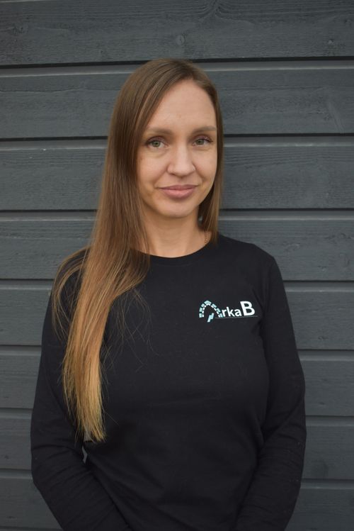 Woman with long brown hair wearing a black shirt with a logo standing against a dark wooden plank wall.