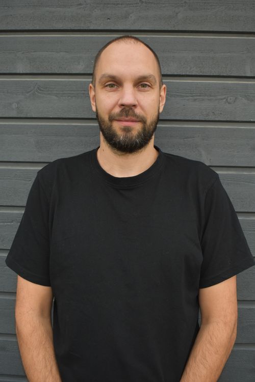 Man with short beard and black t-shirt standing in front of a gray wooden wall, looking at the camera.