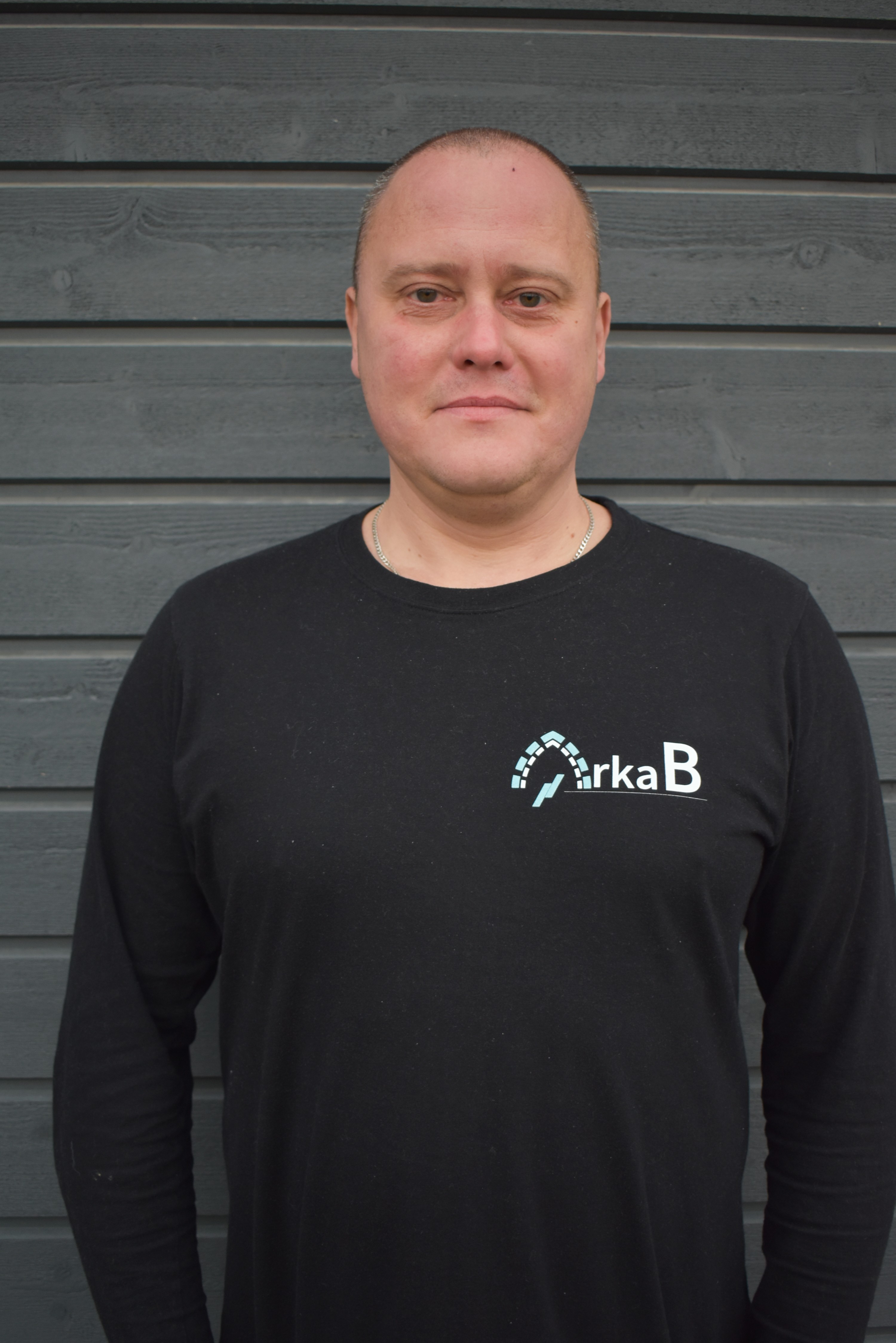 Man with short hair wearing a black shirt with a logo standing against a gray wooden wall.