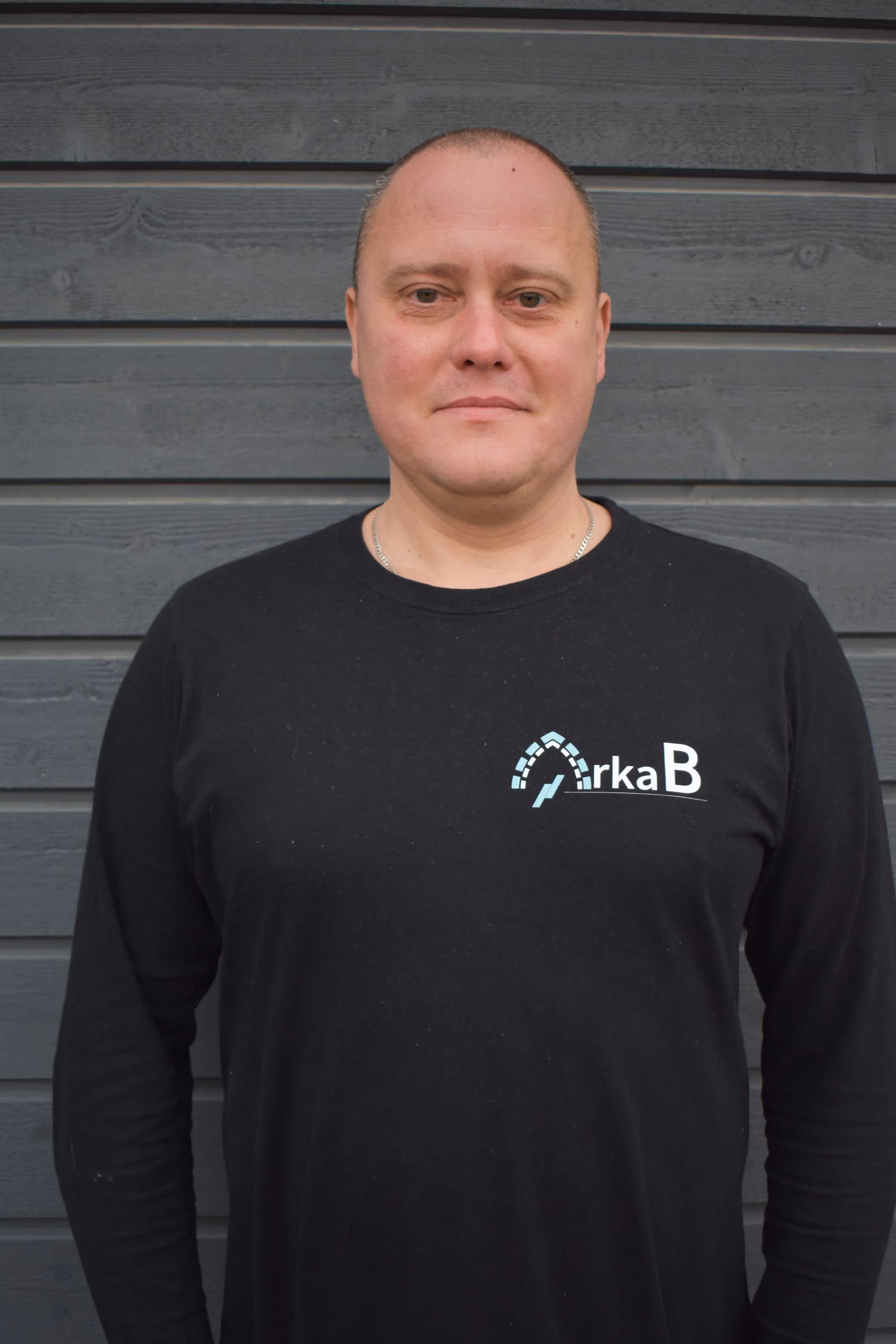Man with short hair wearing a black shirt with a logo standing against a gray wooden wall.