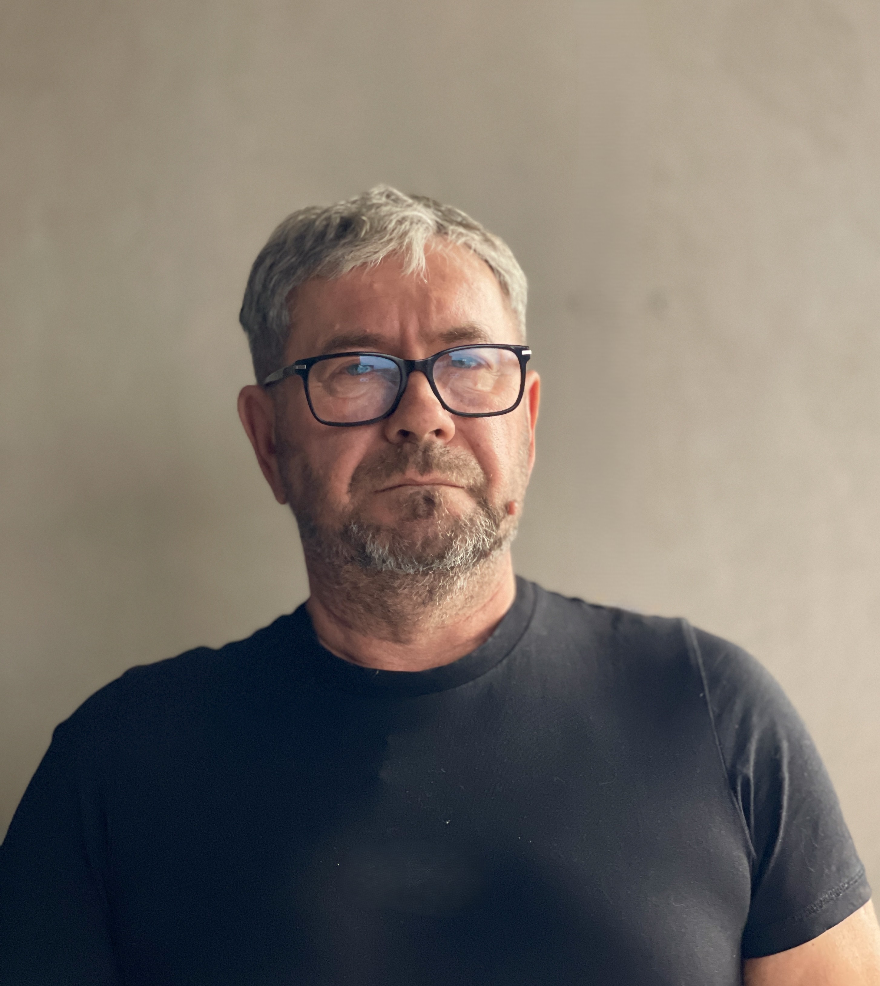 Middle-aged man with gray hair, glasses, and a short beard wearing a black t-shirt against a plain neutral background.
