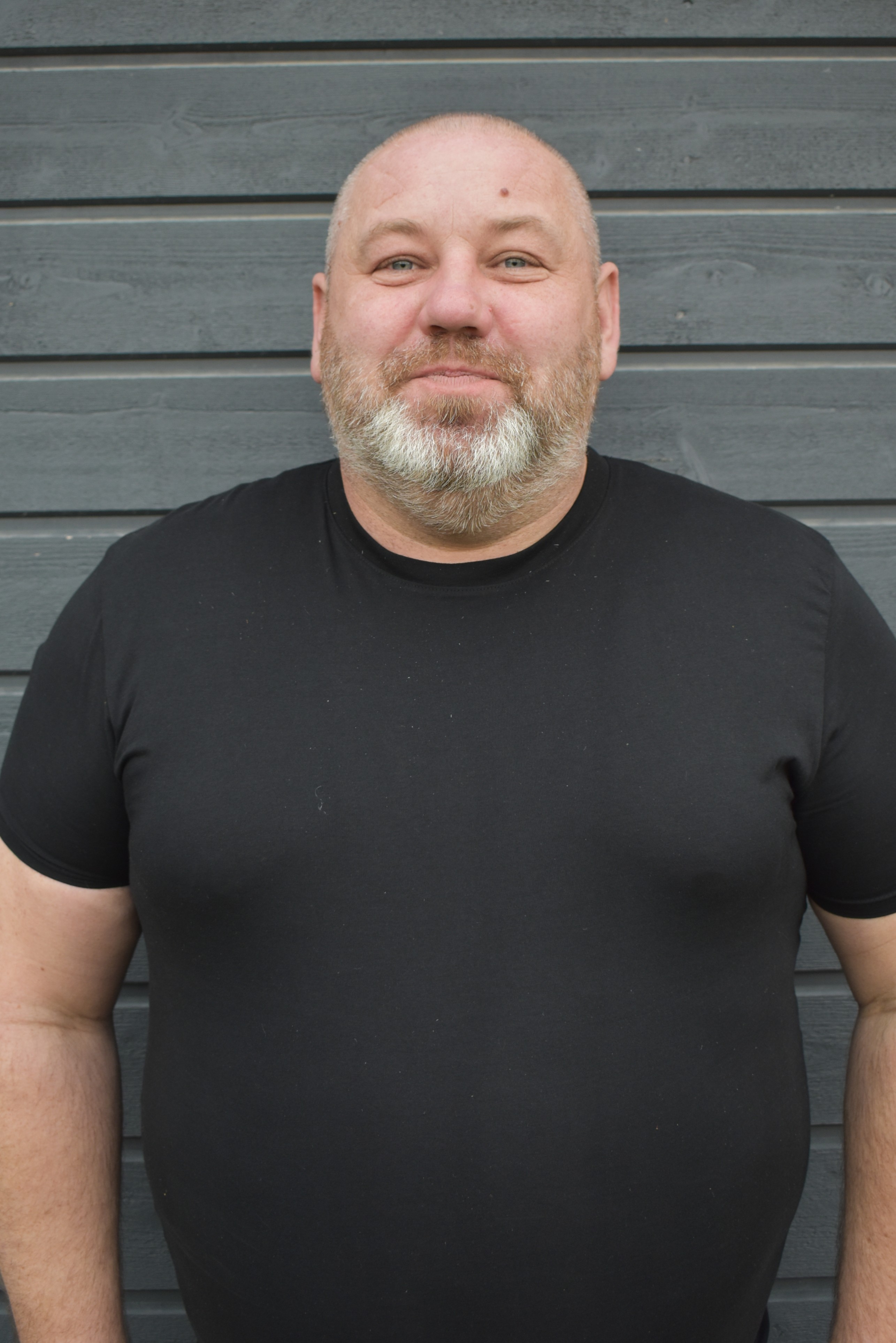Bald man with a gray beard wearing a black t-shirt standing in front of a gray wooden wall.