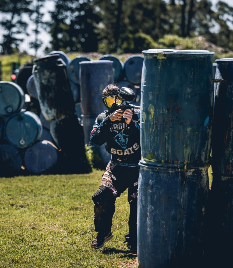 A paintball player crouches behind blue barrels, aiming and prepared to shoot. Bright daylight enhances the scene on a green field at a paintball field, with trees in the background. The subject's gear features a logo, suggesting participation in competitive paintball games.