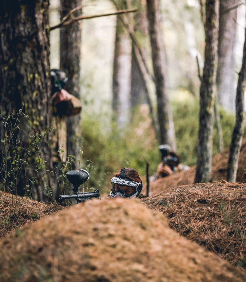A paintball field in a forested area, featuring trees and covered terrain. Players in camouflage can be spotted behind trees, preparing for paintball games. The scene showcases the natural environment of Central Coast paintball and PPK Paintball Kulnura.