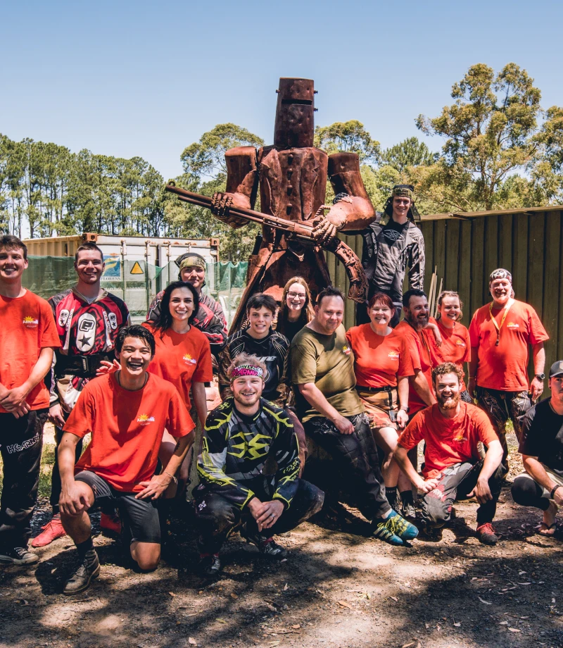 A large group of paintball players in orange shirts poses for a photo outdoors, standing in front of a towering metal sculpture resembling a warrior. The setting is surrounded by trees and paintball structures, creating a playful atmosphere for Central Coast paintball games.