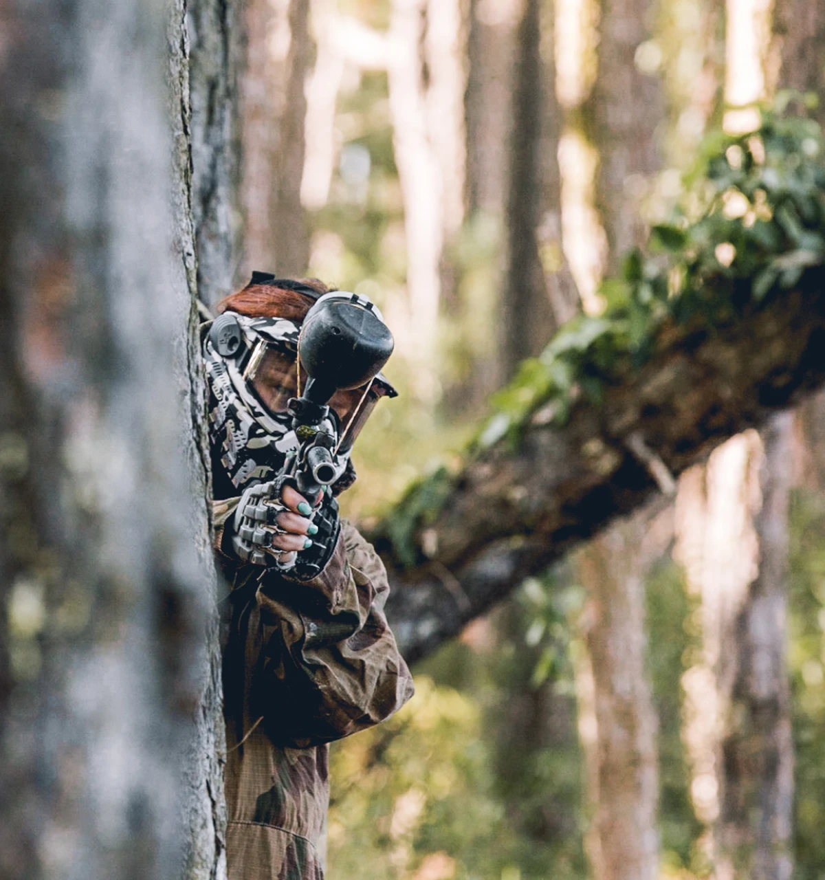 A player in camouflage gear hiding behind a tree, aiming a paintball gun in a lush forest setting. This scene captures the excitement of paintball games at a Central Coast paintball field, highlighting strategy and adventure.