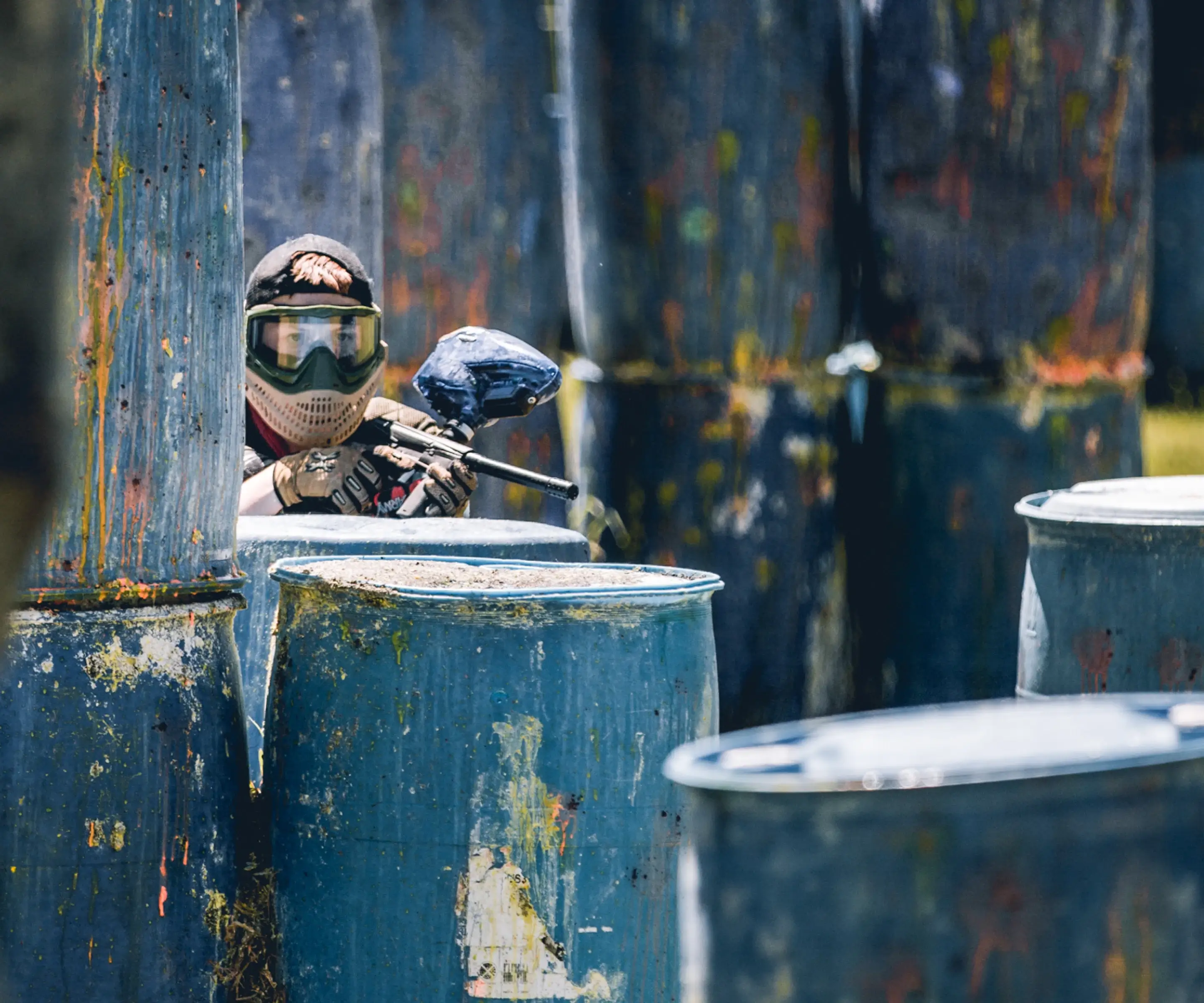 A player in a camouflage outfit and protective gear crouches behind blue barrels on a paintball field, aiming a paintball marker. The vibrant colors and worn surfaces of the barrels highlight the excitement of paintball games at Central Coast paintball.