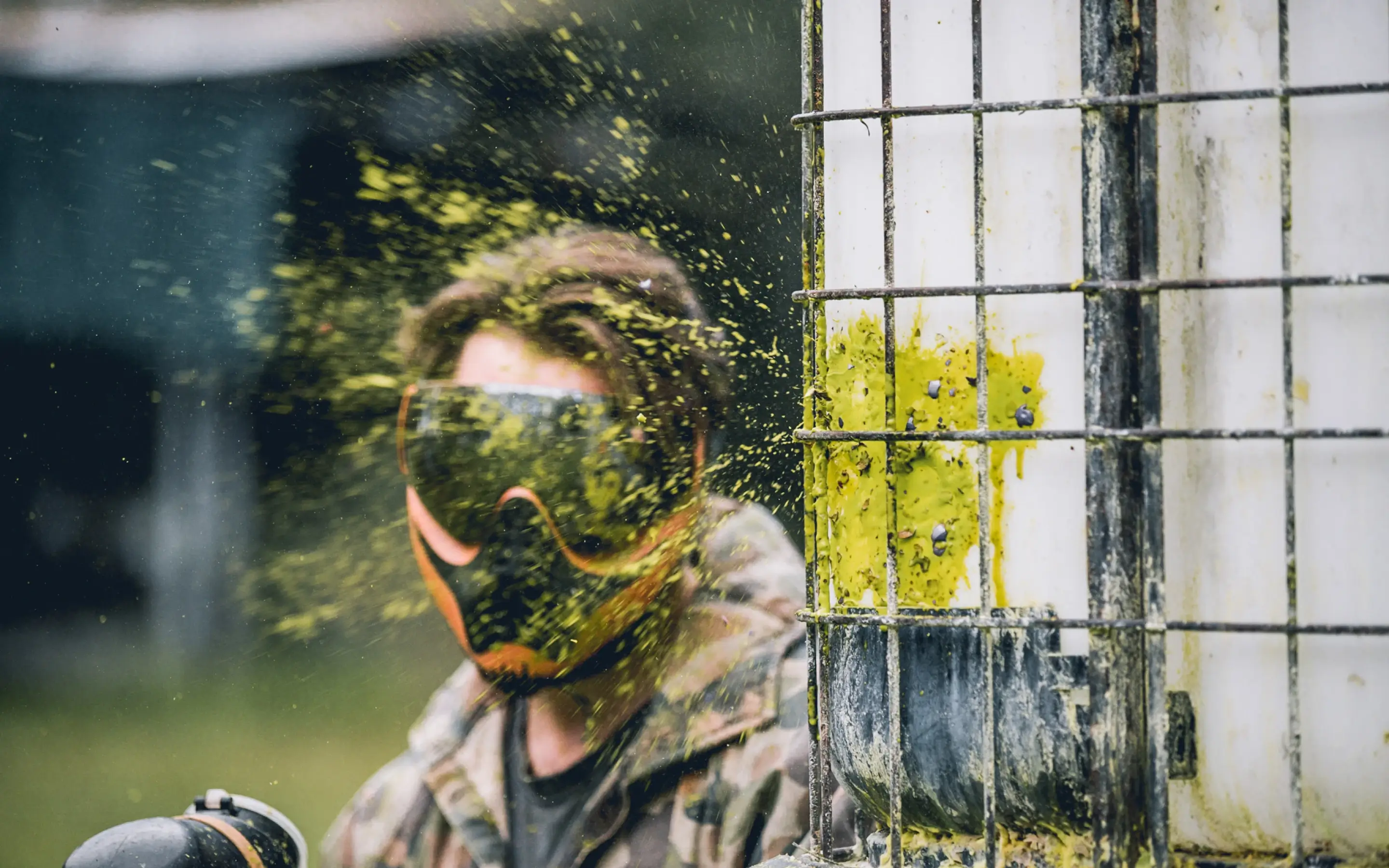 A person wearing a protective mask and camouflage clothing stands near a shooting target, with paint splatter visible in vibrant yellow, indicating a paintball competition. The background features blurred green foliage.