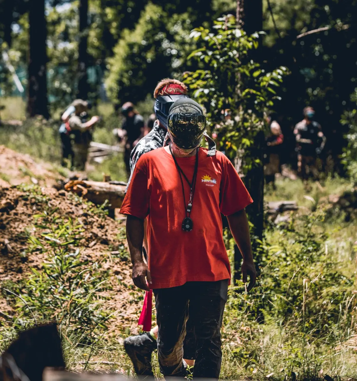 A group of people walking through a wooded area. In the foreground, a man in a red shirt and mask is leading, while others are visible in the background, surrounded by greenery and sunlight filtering through the trees.