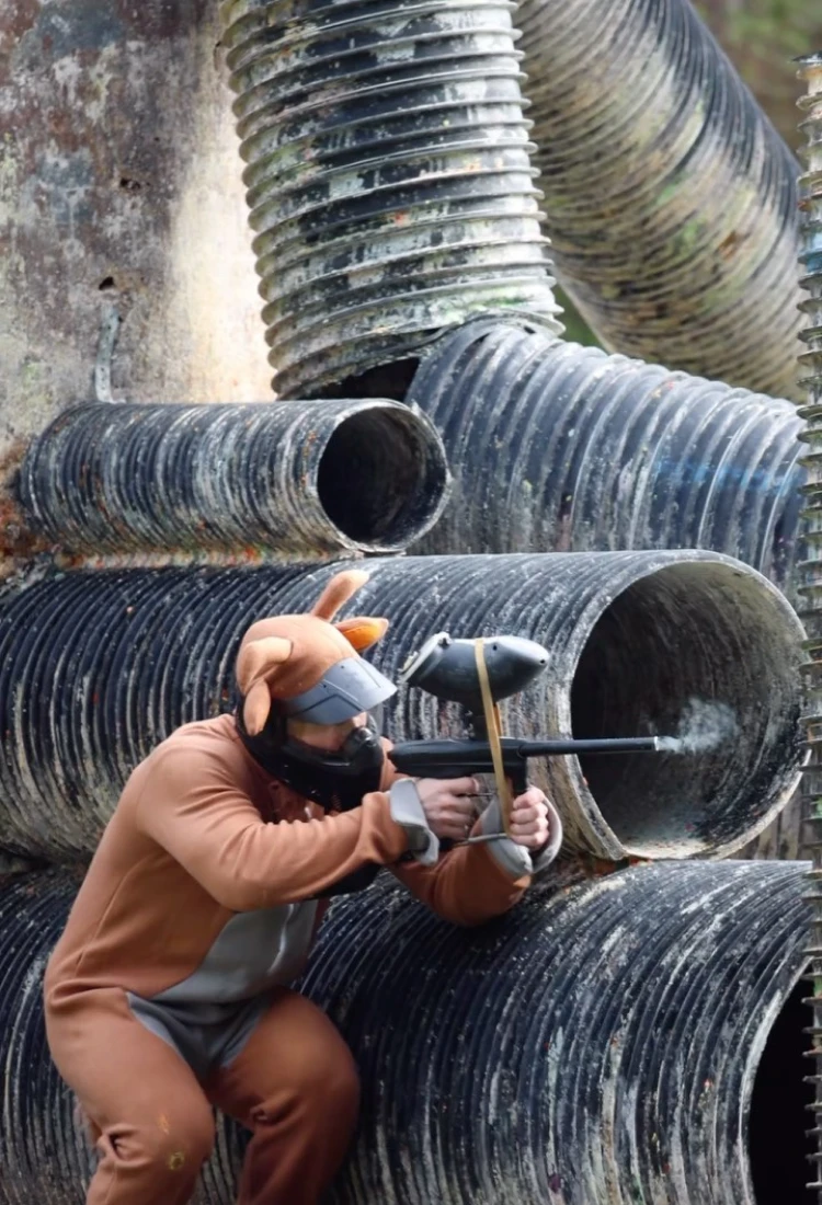 A person dressed in a brown animal costume crouches behind large stacked black plastic pipes, aiming a paintball marker at a target. The setting is an outdoor paintball arena with greenery in the background.