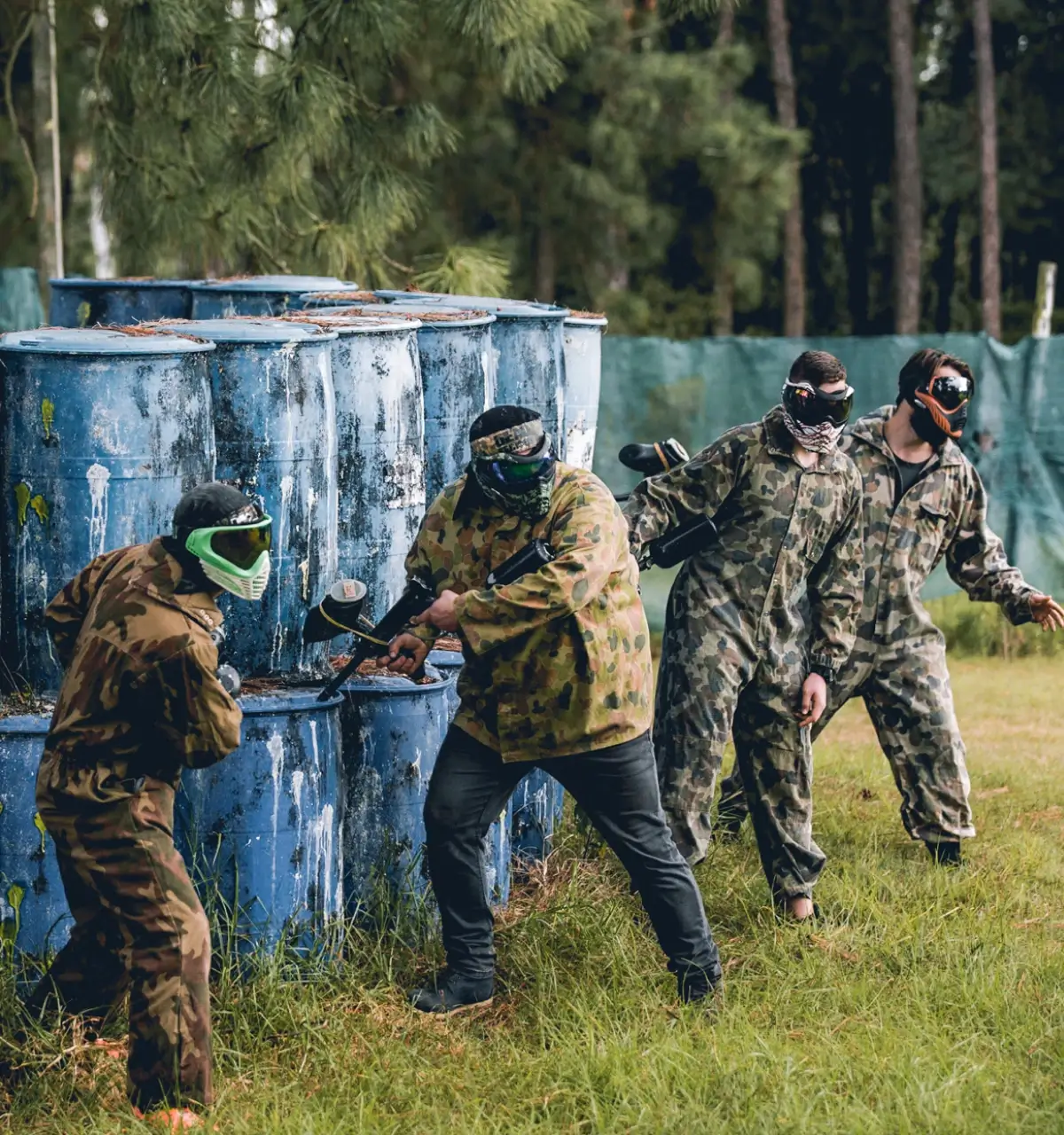 A group of four people in camouflage clothing and protective masks participate in a paintball match. They crouch beside blue barrels in a wooded area, preparing to strategize or engage in the game.