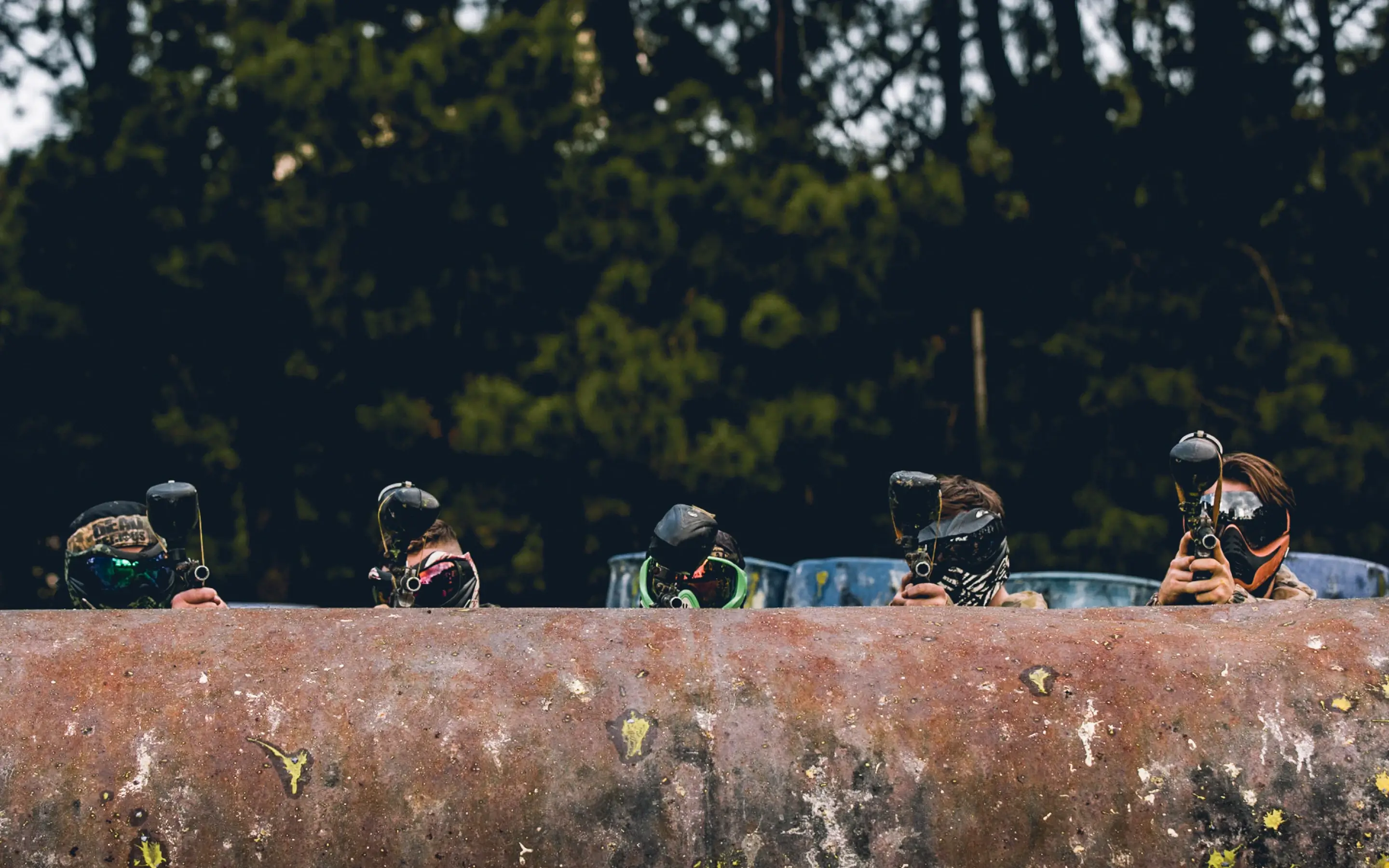 A group of players wearing masks and protective gear, peeking over a rustic barrier during a paintball game in a wooded area.
