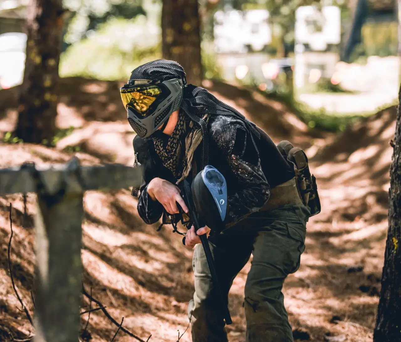 A person wearing a paintball mask and tactical clothing crouches behind a wooden barrier in a forest setting, holding a paintball marker, with sunlight filtering through the trees.