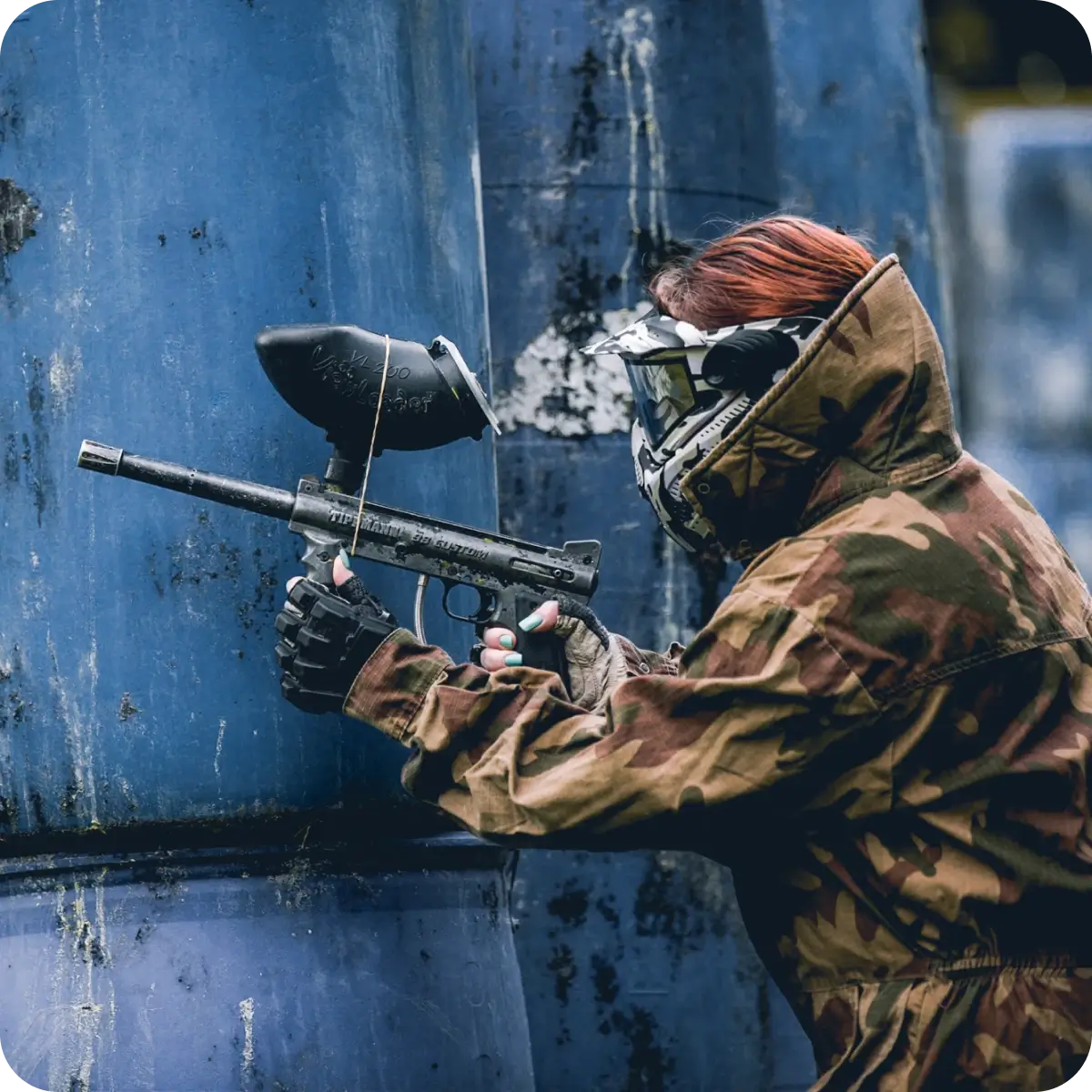 A person in a camouflage outfit and protective mask aims a paintball gun while crouching near a blue structure, showcasing focus and readiness in a competitive paintball setting.