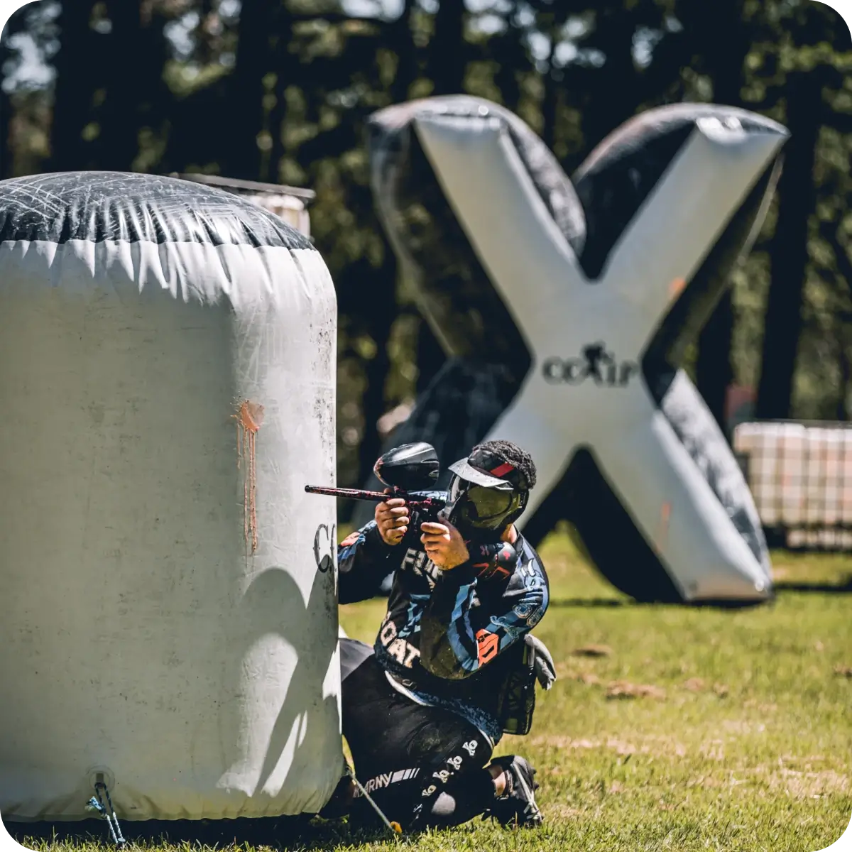 A player kneeling behind a large inflatable barricade in a paintball arena, aiming a marker. Behind him, a large inflatable obstacle shaped like an 'X' towers over the grassy field.