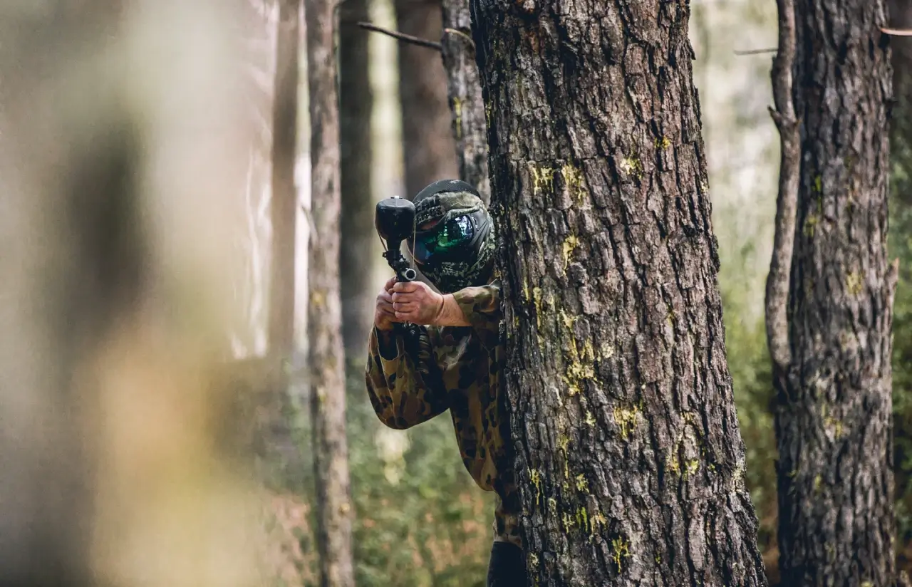 A player in camouflage clothing crouches behind a thick tree in a wooded area, aiming a paintball marker. The sunlight filters through the trees, creating a dappled light effect on the forest floor.
