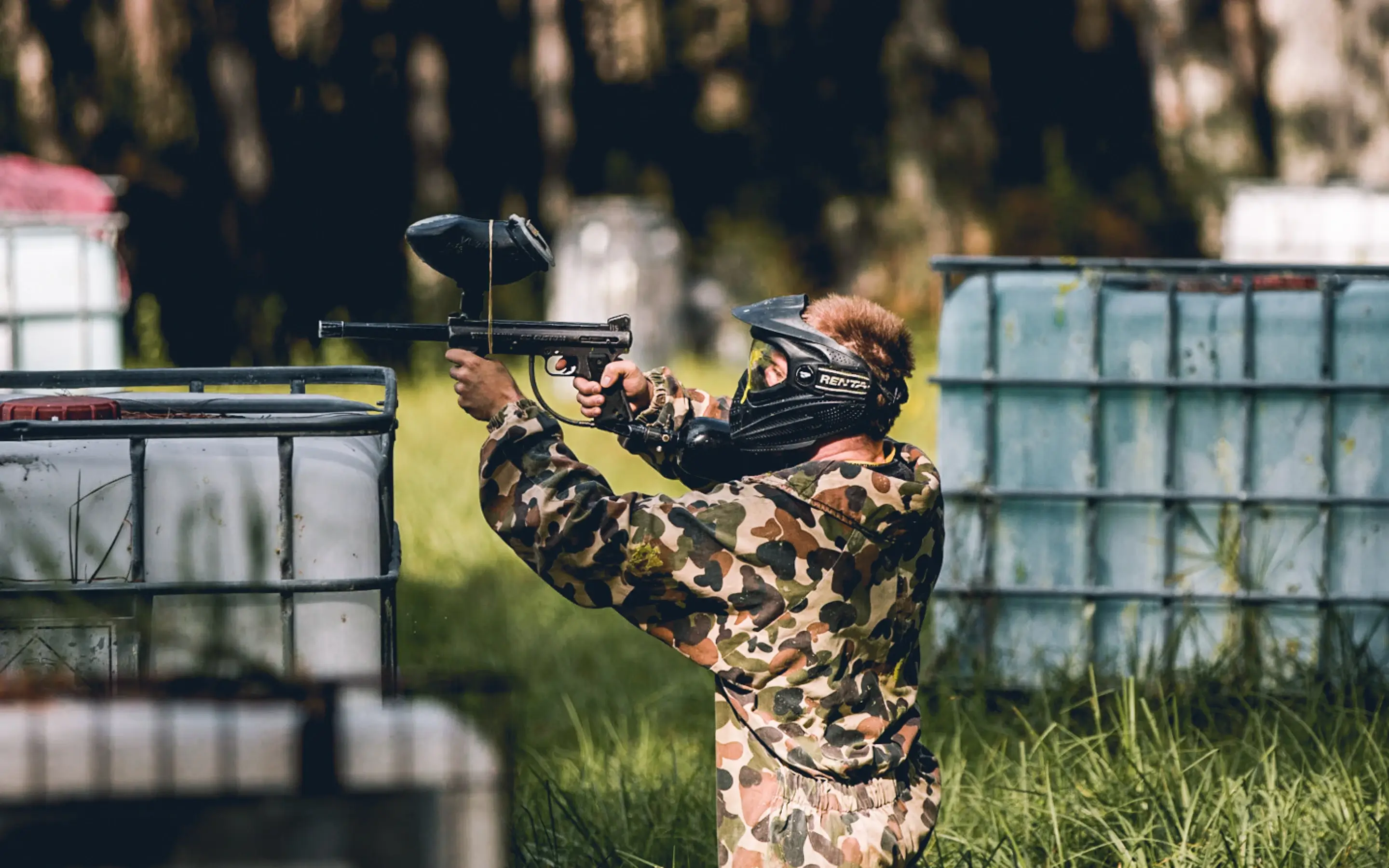 A person in camouflage attire wearing a mask is aiming a paintball gun while crouching behind large, plastic containers. The scene is set in a green, outdoor area with trees in the background.