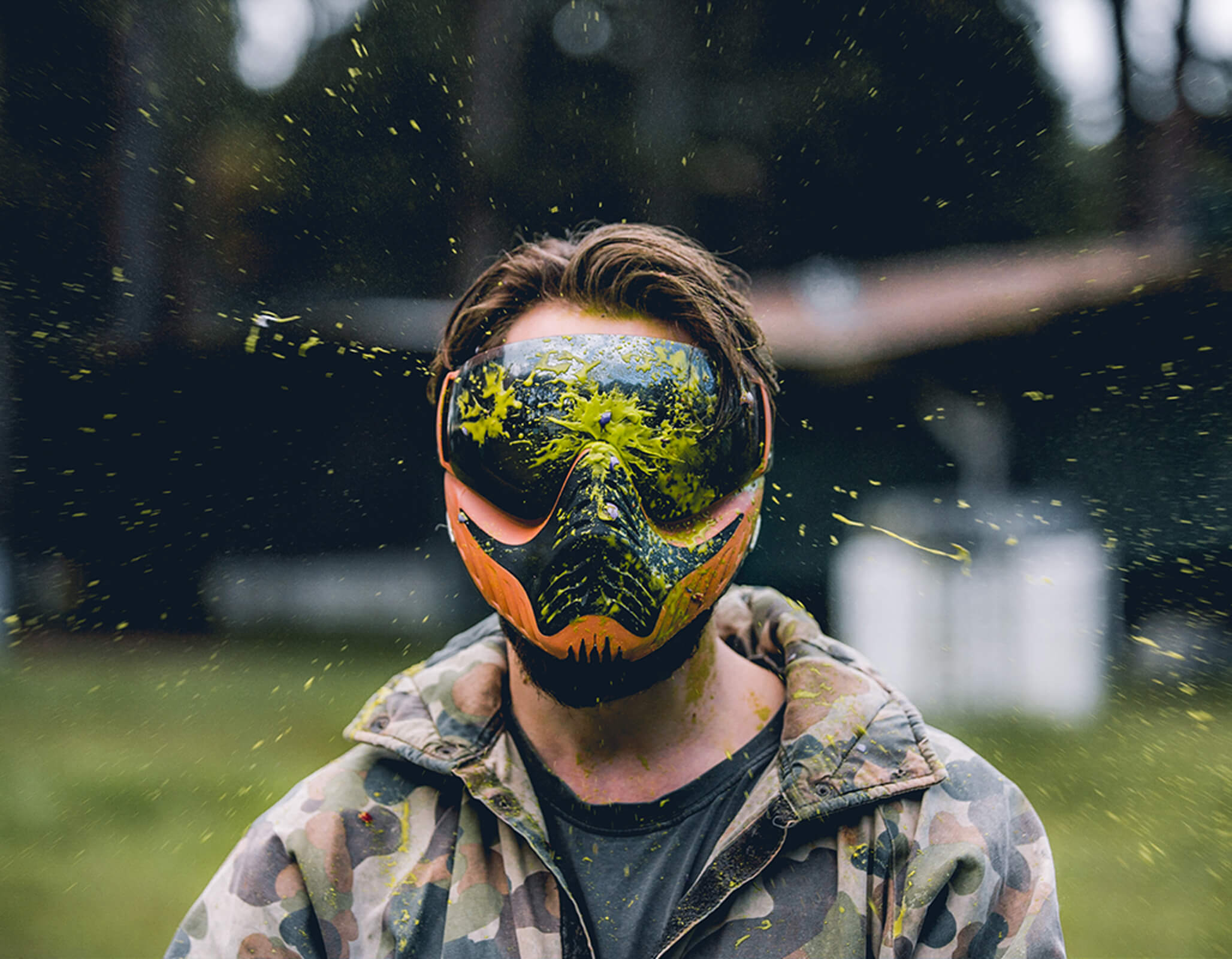 A person wearing a colorful paintball mask that features a striking design, with vibrant splashes of paint surrounding them. Dressed in a camouflage jacket, they stand confidently, ready for action at a Central Coast paintball field.