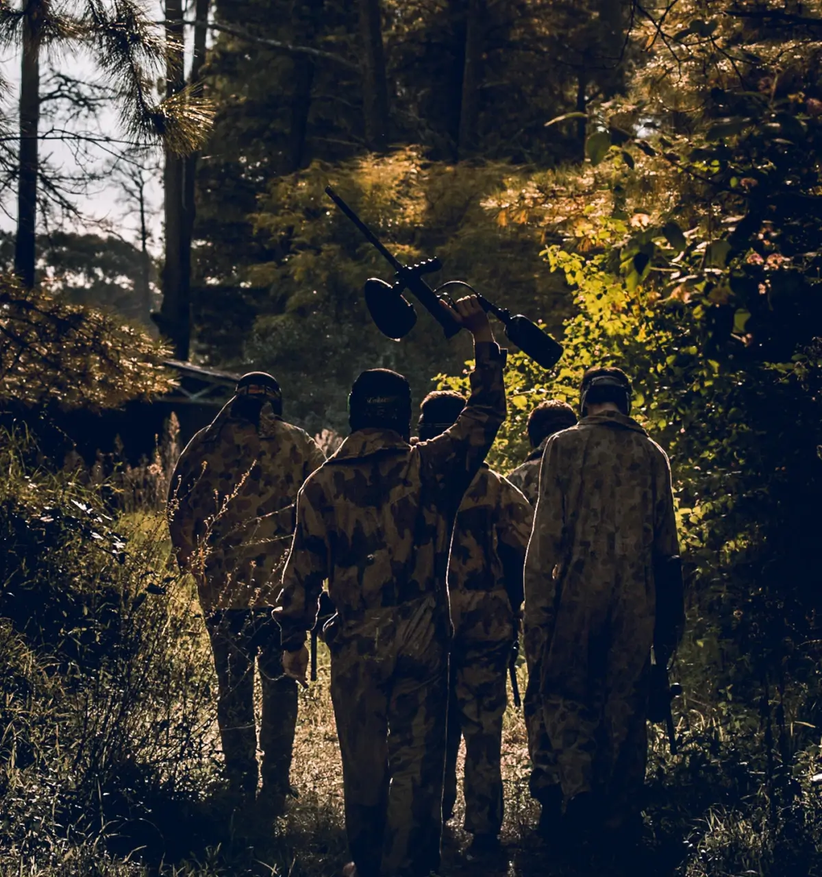 A group of four people in camouflage clothing and protective masks participate in a paintball match. They crouch beside blue barrels in a wooded area, preparing to strategize or engage in the game.