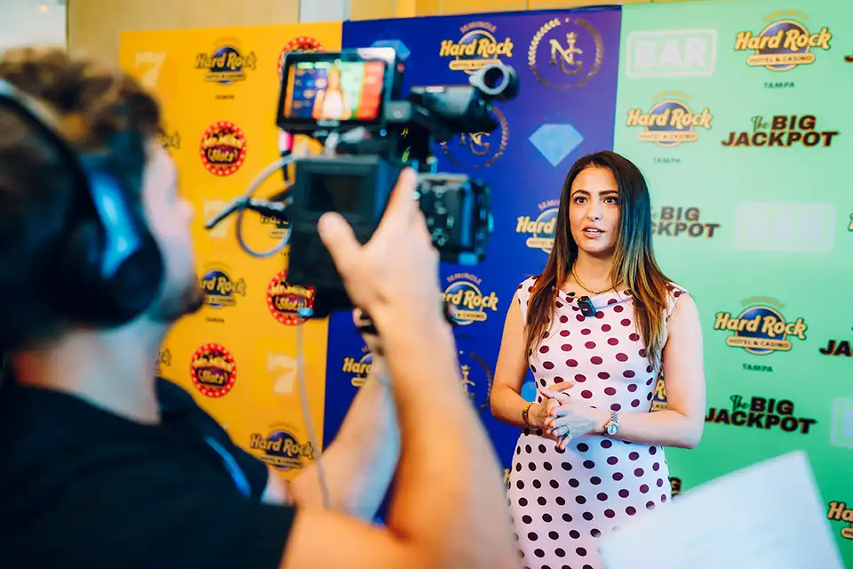 Woman in a white dress with red polka dots speaking on camera in front of a colorful Hard Rock and casino-themed backdrop.