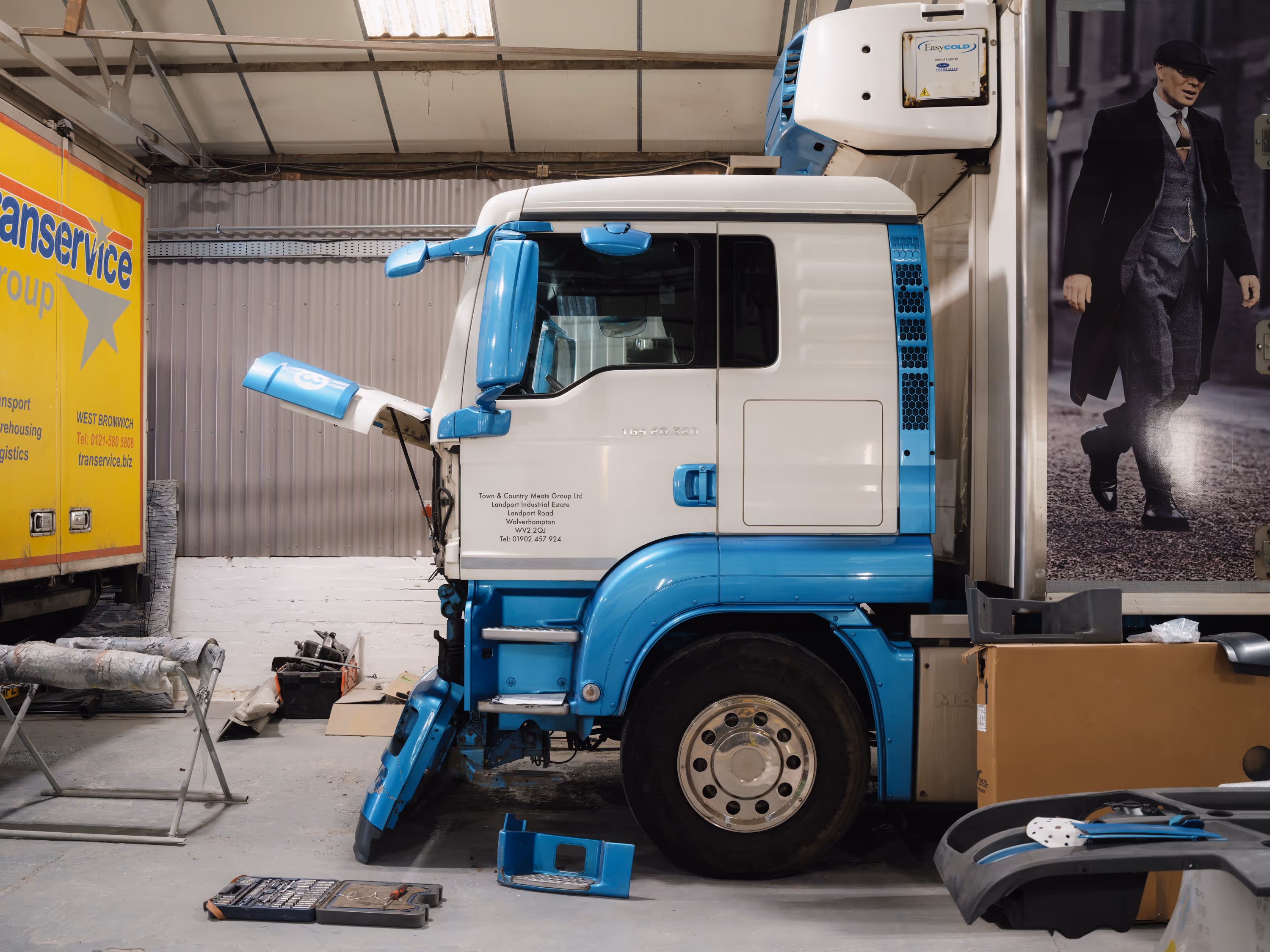 Side view of a blue and white semi-truck cab in a garage with parts removed and repair tools on the floor.